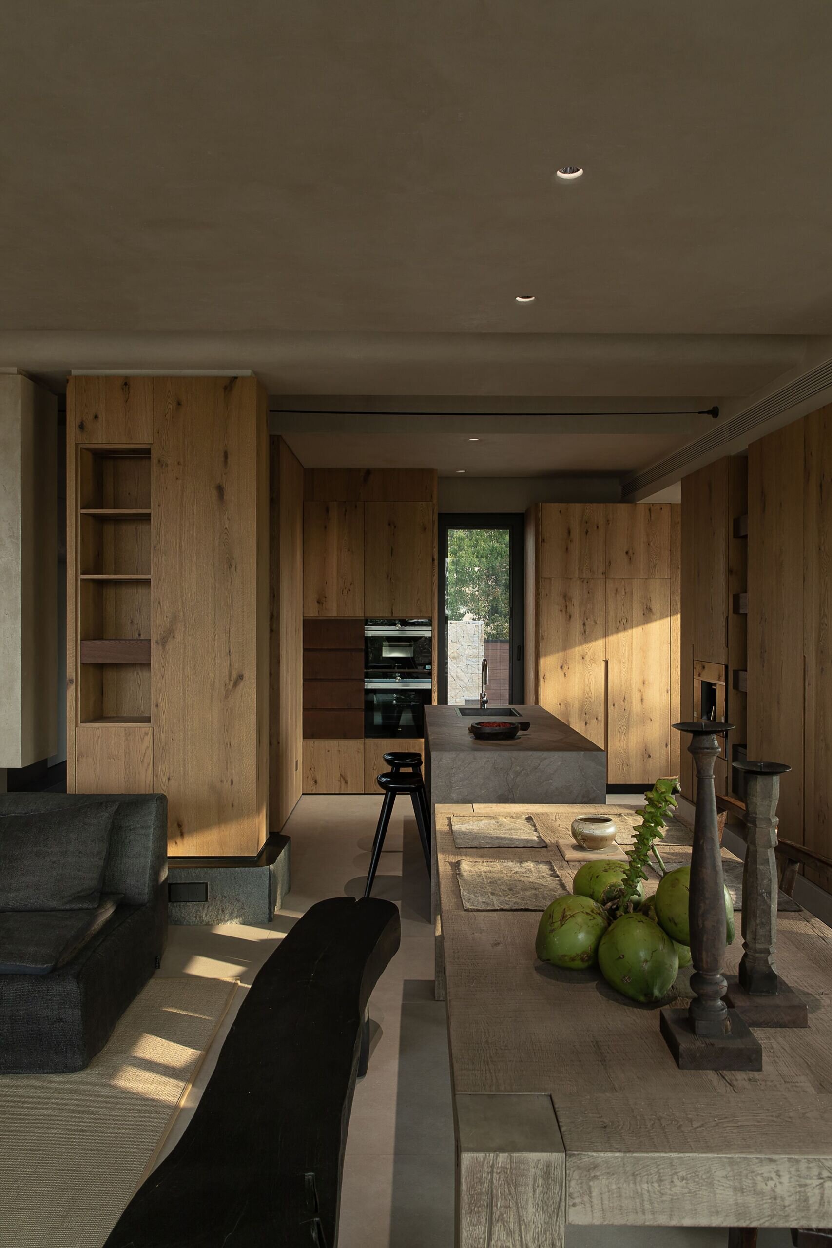 This kitchen and dining space feels richly grounded in natural textures, where raw wood cabinetry wraps the room in warmth and calm. The stone island adds sculptural contrast, its soft, matte finish catching the afternoon light just right, while a long dining table anchors the scene with rustic charm.