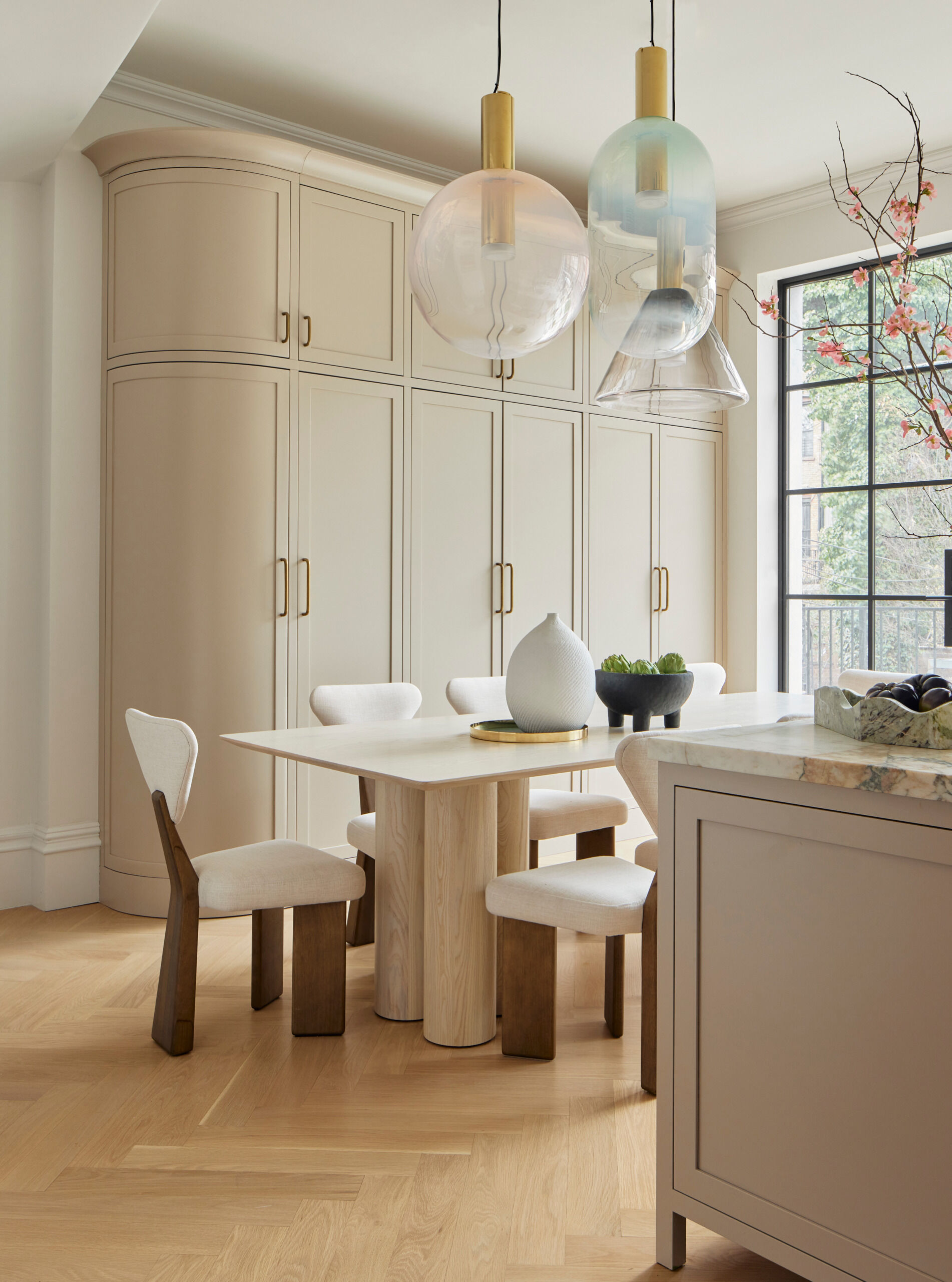 The kitchen is grounded by Calacatta Vagli Viola marble countertops and an island painted in warm mushroom tones. Custom cabinetry and a curved pantry bring softness to the architecture, while a trio of hand-blown glass pendants hang above a pale oak dining table. It’s an inviting space that balances simplicity with elegance, ready for casual meals or larger gatherings.