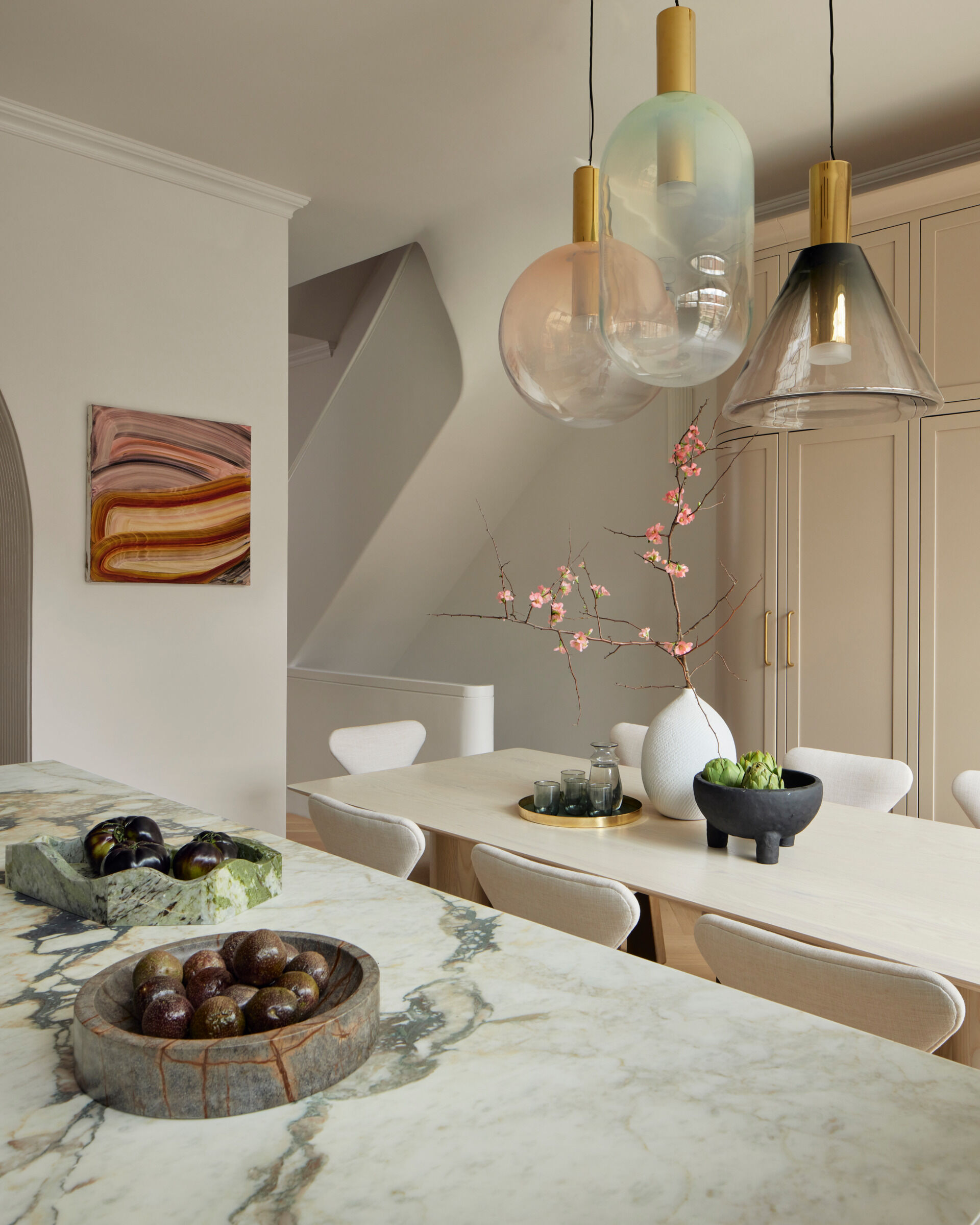 The kitchen is grounded by Calacatta Vagli Viola marble countertops and an island painted in warm mushroom tones. Custom cabinetry and a curved pantry bring softness to the architecture, while a trio of hand-blown glass pendants hang above a pale oak dining table. It’s an inviting space that balances simplicity with elegance, ready for casual meals or larger gatherings.