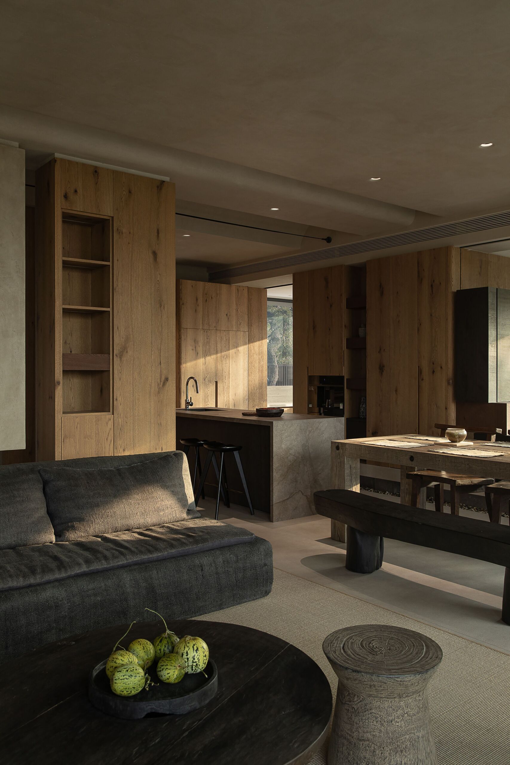 This kitchen and dining space feels richly grounded in natural textures, where raw wood cabinetry wraps the room in warmth and calm. The stone island adds sculptural contrast, its soft, matte finish catching the afternoon light just right, while a long dining table anchors the scene with rustic charm.