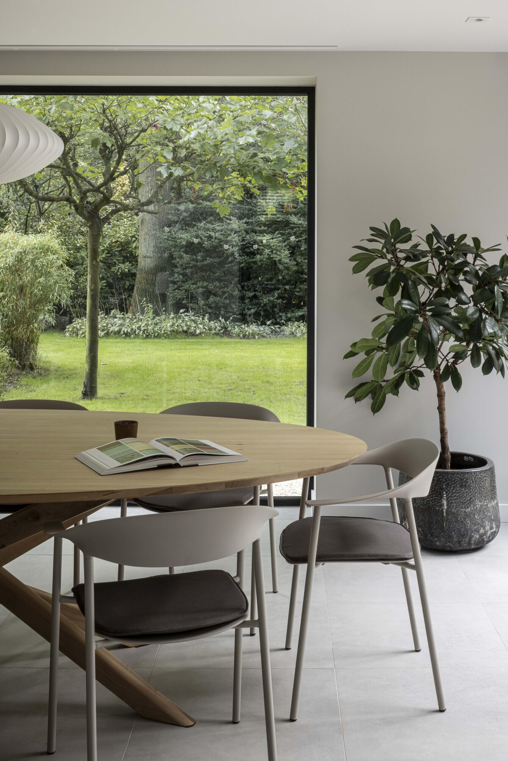 This dining area offers a peaceful blend of natural light, soft tones, and organic textures. A large picture window frames the lush green garden outside, turning the view into a living artwork that changes with the seasons.  The oval wooden table features a sculptural base that adds quiet interest without overwhelming the space. Surrounding it, minimalist chairs in pale gray with dark cushions maintain the room’s calm, understated palette. A single open book rests on the table, adding a sense of lived-in warmth and quiet reflection.  To the side, a potted indoor tree in a textured charcoal planter brings nature indoors and balances the strong vertical line of the window. Altogether, this space feels serene and grounded, ideal for both everyday meals and thoughtful moments.