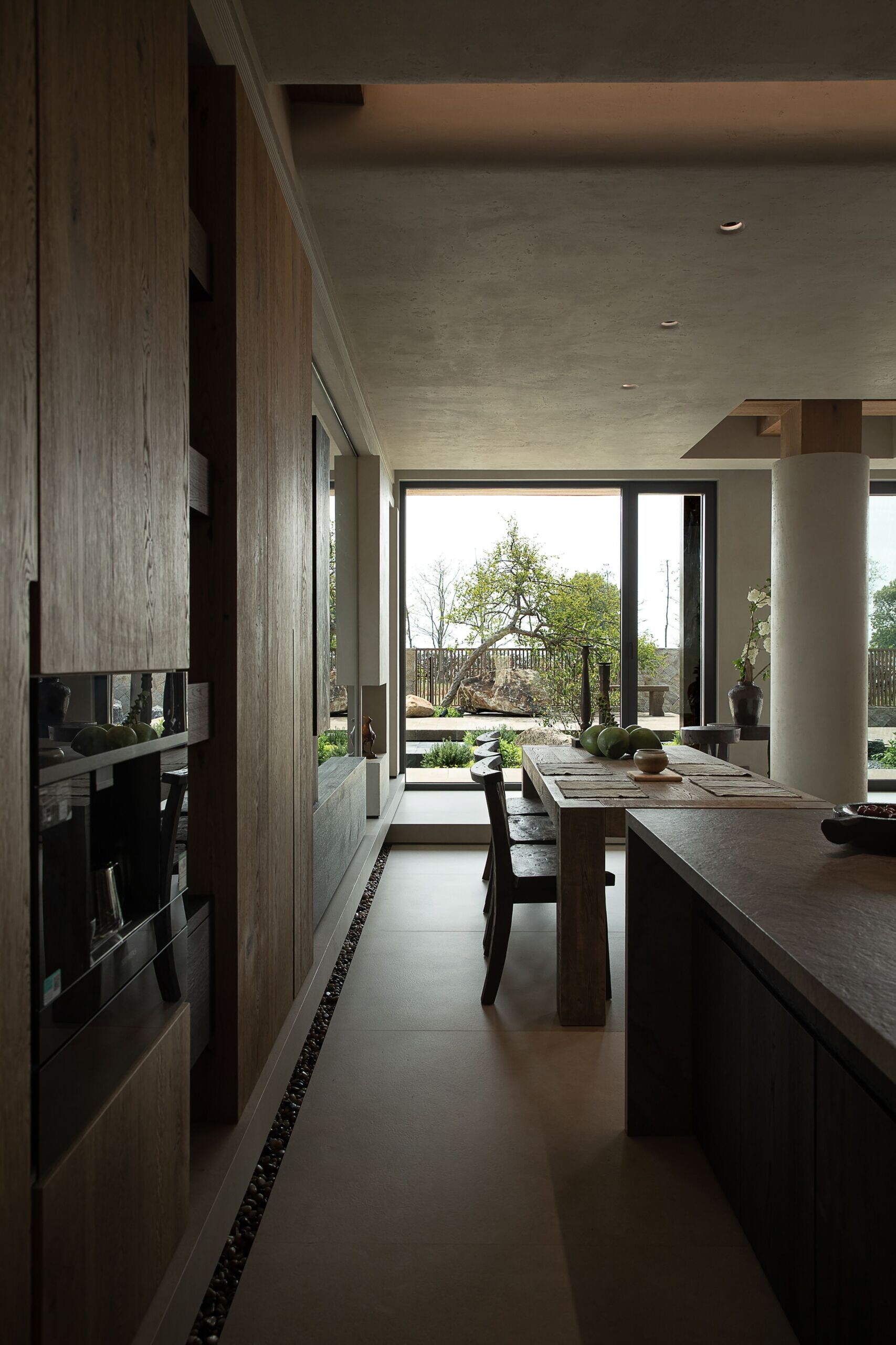 This kitchen and dining space feels richly grounded in natural textures, where raw wood cabinetry wraps the room in warmth and calm. The stone island adds sculptural contrast, its soft, matte finish catching the afternoon light just right, while a long dining table anchors the scene with rustic charm.
