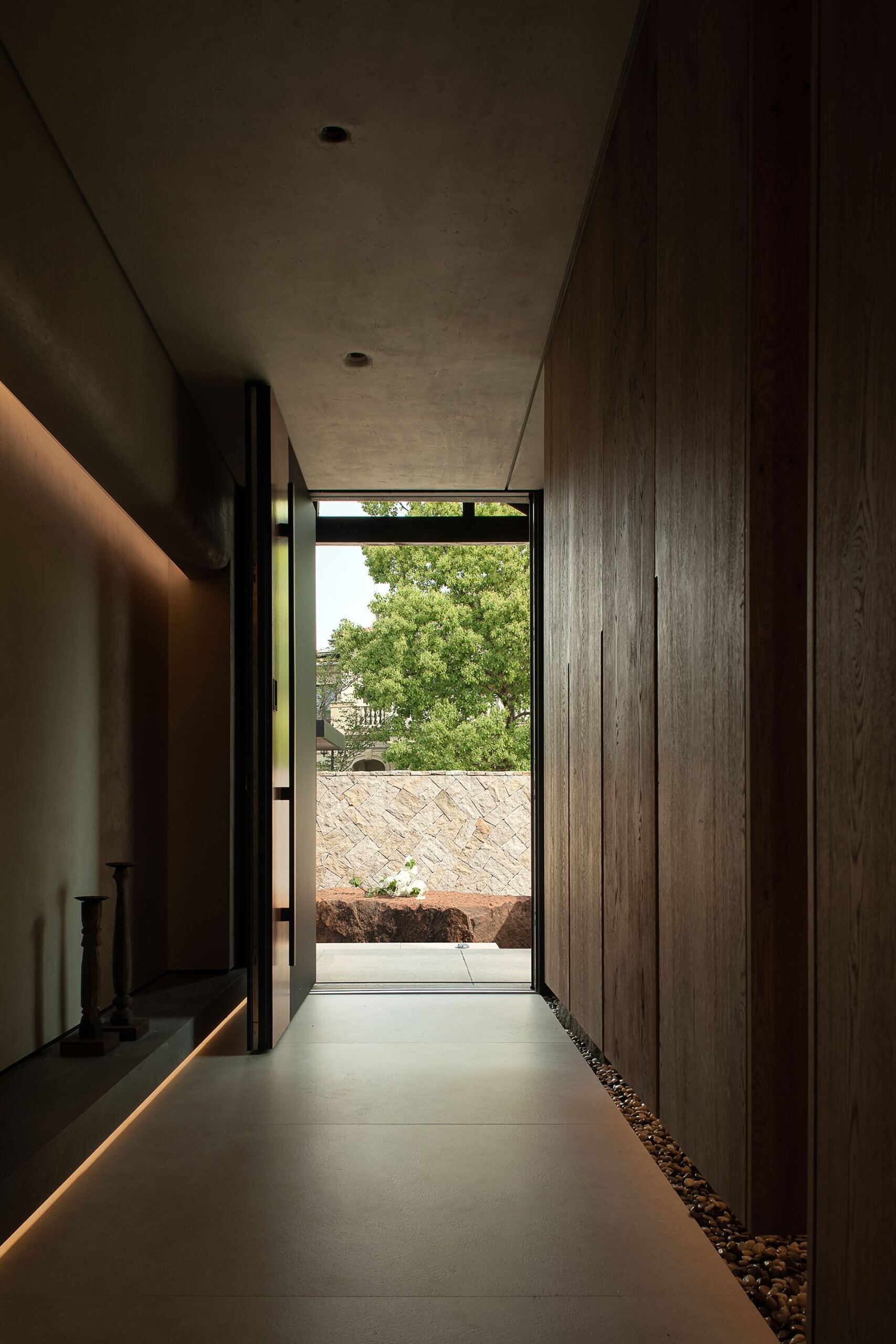 This entryway is all about quiet drama and natural flow. Soft light traces along the floor and walls, gently highlighting the smooth concrete and dark wood surfaces. A narrow strip of river stones tucked along the base adds texture and a subtle connection to the outdoors.
