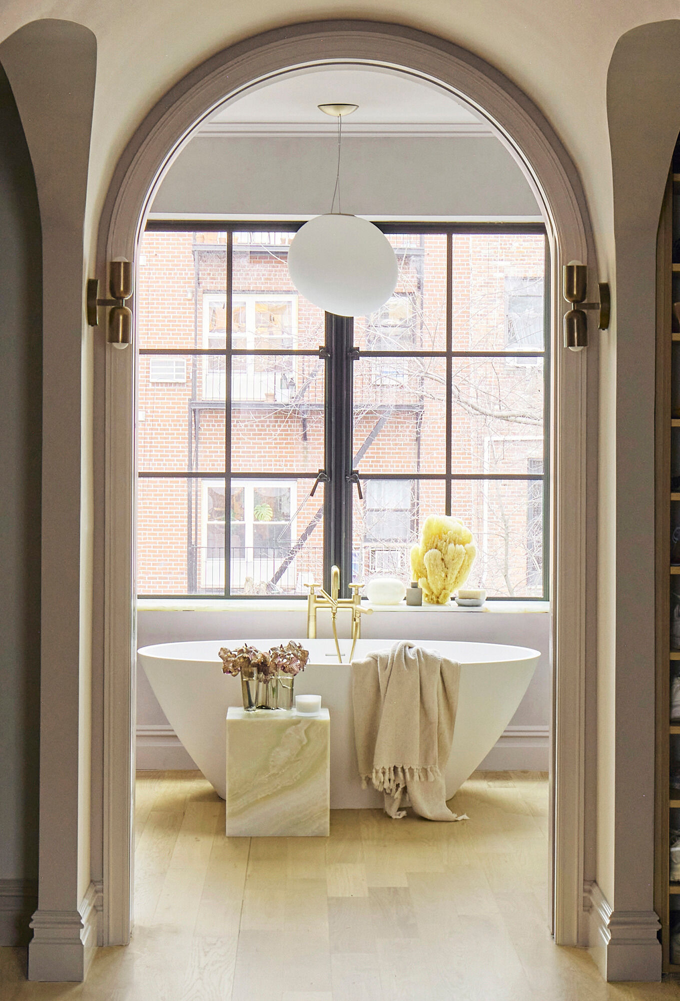This primary bathroom continues the lavender-gray plaster from the bedroom and elevates it with dramatic stone selections. Green onyx and rosa aurora macchia vecchia marble define the shower, while a freestanding tub sits beneath a steel casement window overlooking the garden and the wood vanity is topped with onyx.