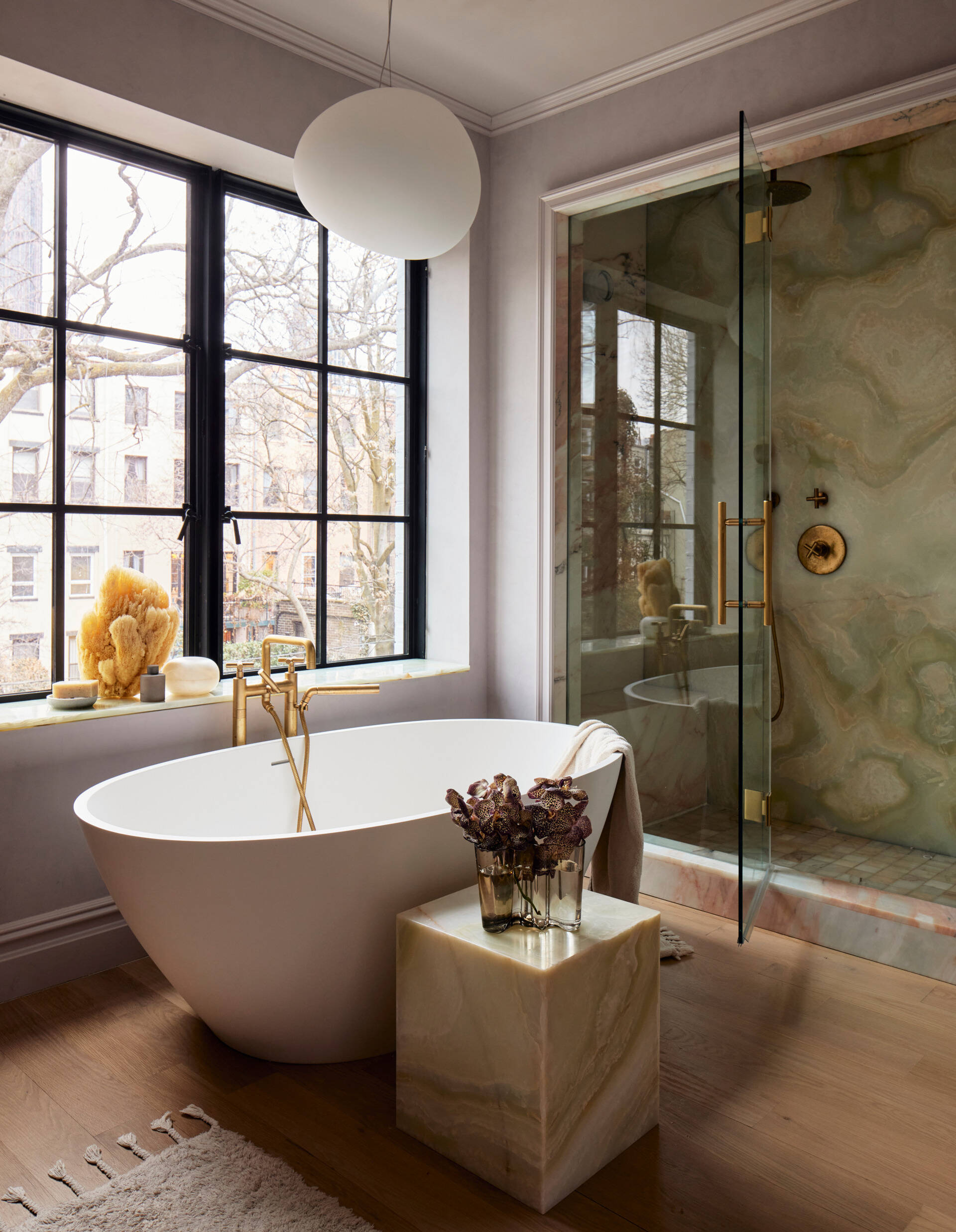 This primary bathroom continues the lavender-gray plaster from the bedroom and elevates it with dramatic stone selections. Green onyx and rosa aurora macchia vecchia marble define the shower, while a freestanding tub sits beneath a steel casement window overlooking the garden and the wood vanity is topped with onyx.
