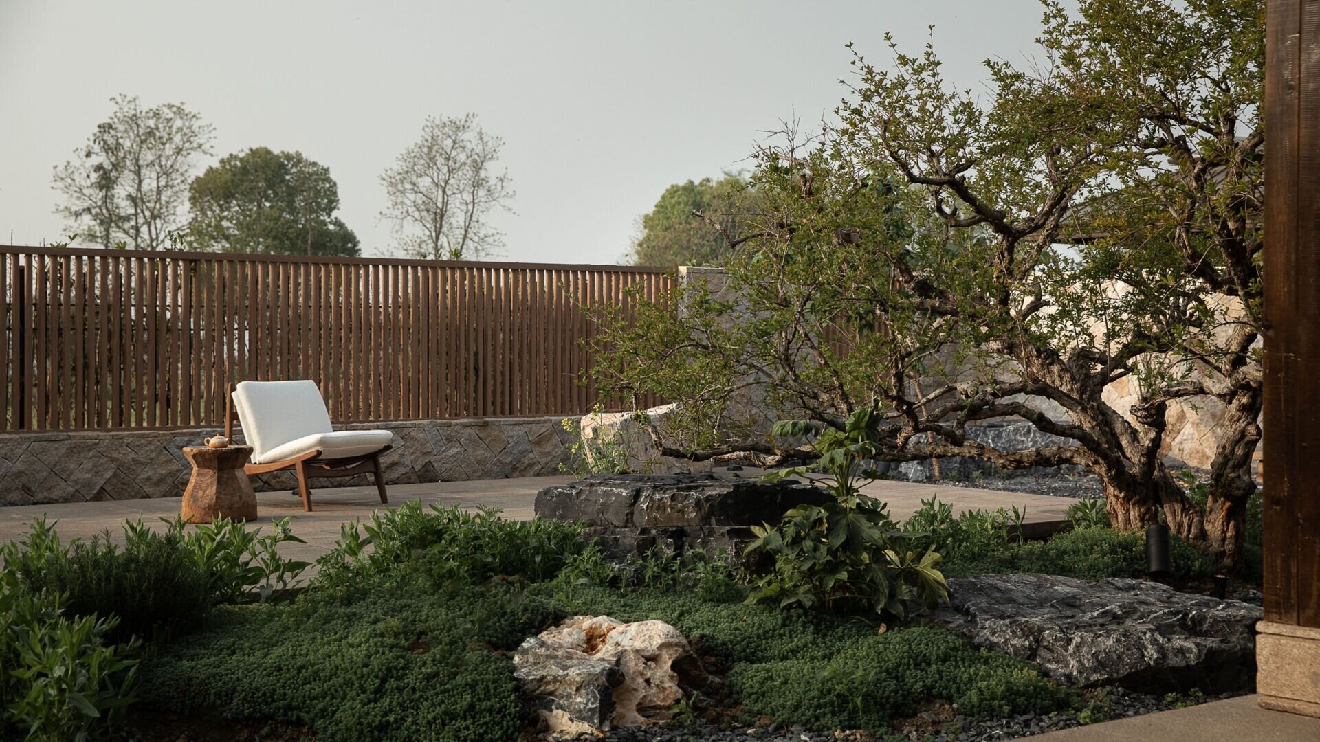 This courtyard expands to include lush green groundcover, softening the base of gnarled tree trunks and polished black stone. A single low chair, paired with a carved wood side table, invites slow mornings with tea or a book. The vertical slat fence adds privacy without closing off the view, letting filtered light pour in while keeping the space peaceful and secluded.