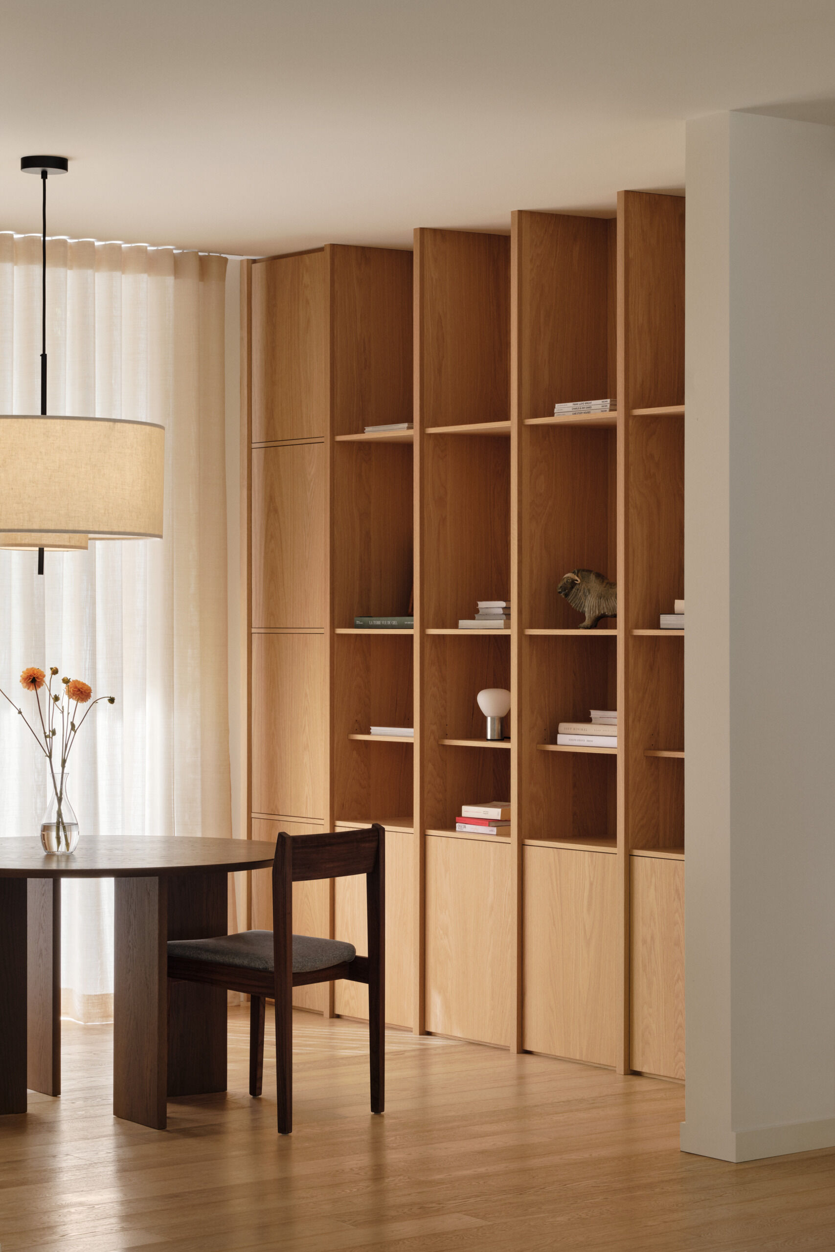 This sunlit home library offers a quiet spot for reading or morning coffee. Oak shelving lines the wall with a calm rhythm, styled simply with books and objects. A dark wood table with matching chairs anchors the space, while sheer curtains filter light, casting a warm, inviting glow.