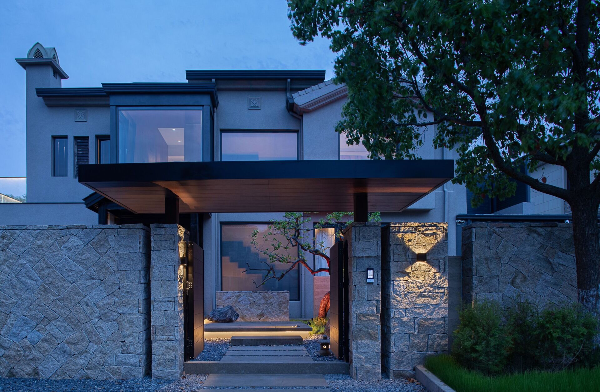 Cool gray stone, soft architectural lighting, and crisp black framing give the entrance to the home a quietly striking presence. The geometric lines feel grounded yet elegant, while warm wood details under the canopy offer a subtle, welcoming contrast.