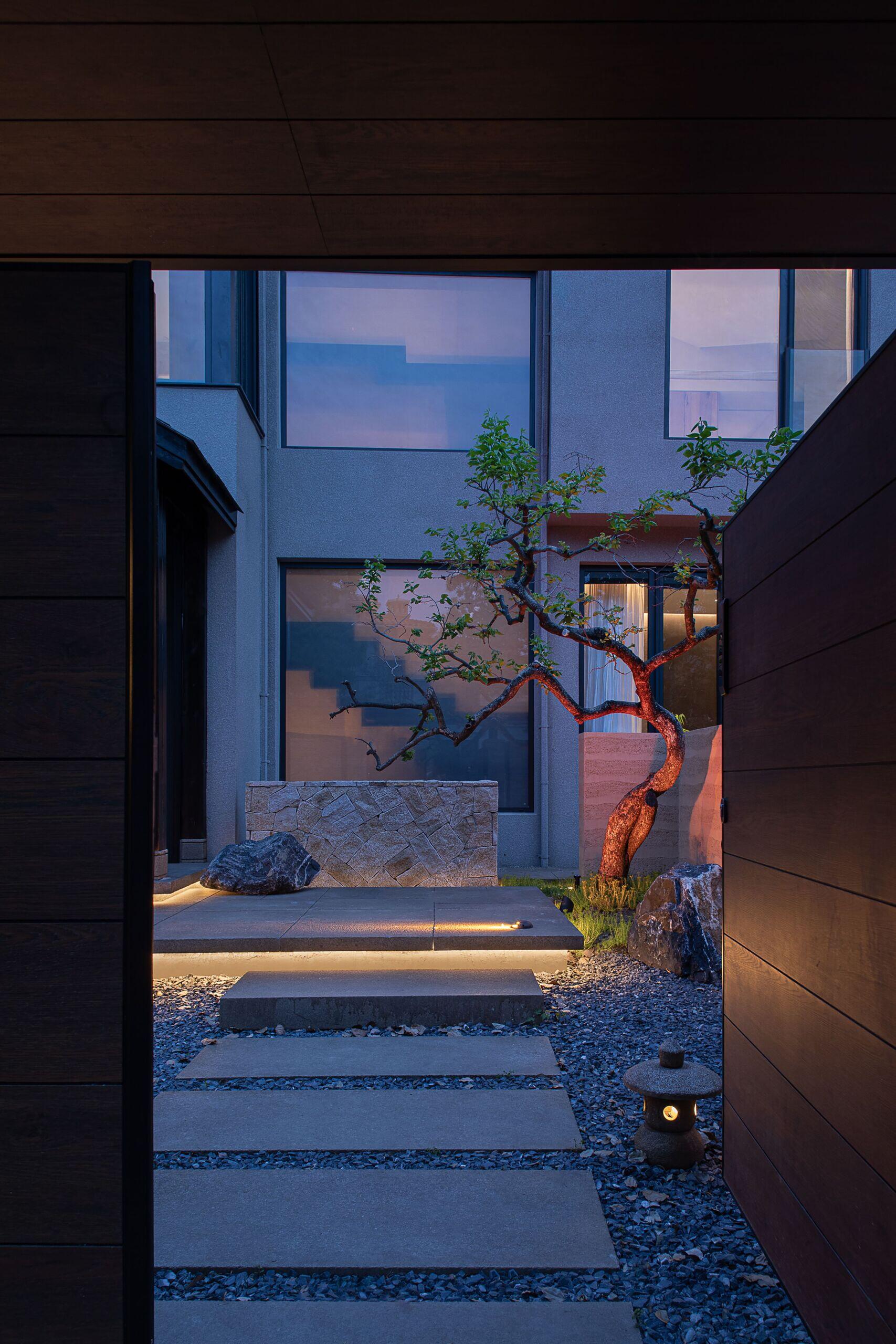 A quiet courtyard draws you in with its serene composition of soft stone, glowing path lights, and a single sculptural tree that feels almost poetic against the dusky sky. Gentle uplighting highlights the tree’s twisted trunk and casts long, peaceful shadows on the textured walls. Underfoot, smooth concrete pavers float over a bed of gravel, creating a soothing rhythm with each step.