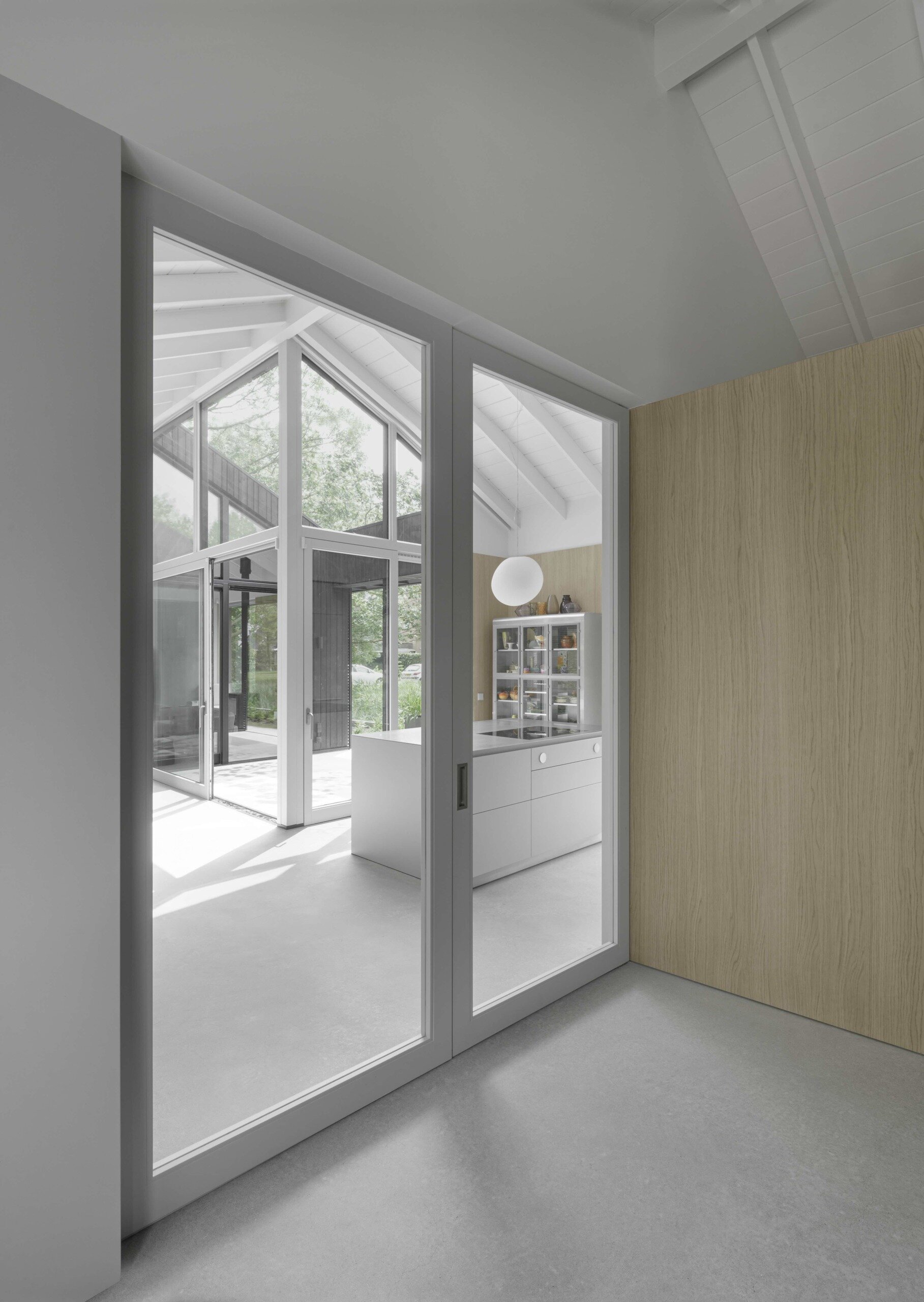 A simple front door opens to a small foyer where a sliding glass door slides back to reveal the home’s heart, inviting natural light and uninterrupted sightlines. Light grey concrete floors stretch throughout the house, grounding the interiors in a calm, neutral tone. Oak wall cladding wraps the rooms in warmth, while the exposed white-painted rafters overhead establish a continuous rhythm and unify the spaces.