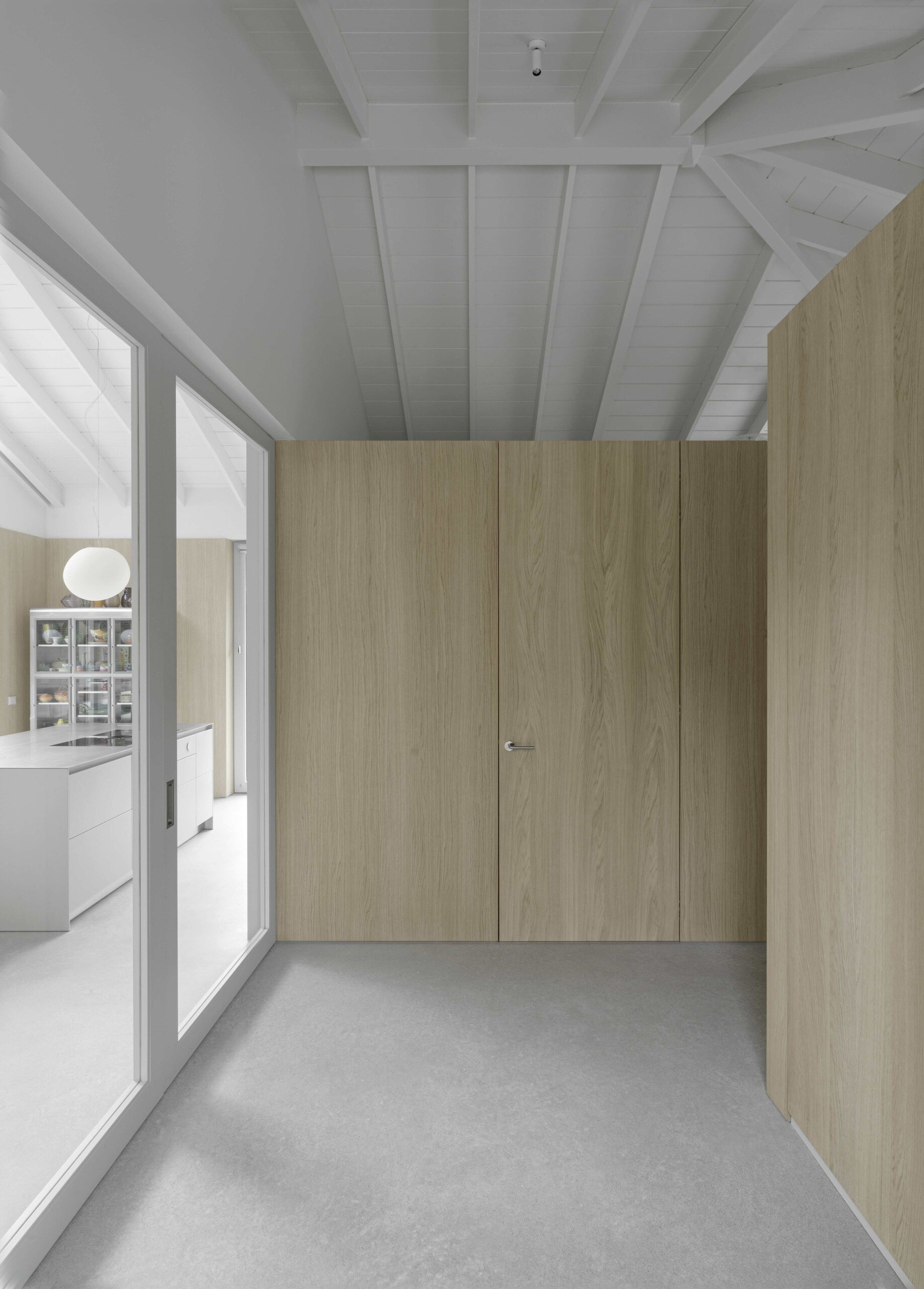A simple front door opens to a small foyer where a sliding glass door slides back to reveal the home’s heart, inviting natural light and uninterrupted sightlines. Light grey concrete floors stretch throughout the house, grounding the interiors in a calm, neutral tone. Oak wall cladding wraps the rooms in warmth, while the exposed white-painted rafters overhead establish a continuous rhythm and unify the spaces.
