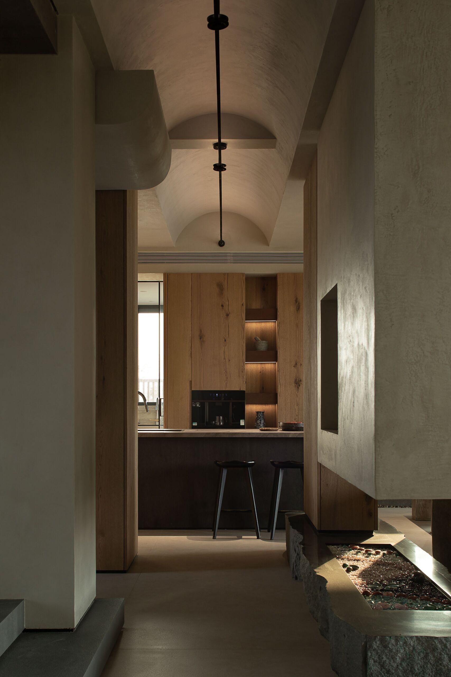 Softly lit and full of texture, this view toward the kitchen shows how the arched ceiling adds a sense of volume and calm, while warm wood cabinetry and stone accents create a grounded, natural palette. A slender light fixture runs cleanly overhead, guiding the eye toward the cozy bar seating. Just in front, a unique recessed fireplace with polished stones and flickering flames adds unexpected warmth, both visual and literal, to this elegant transitional space.