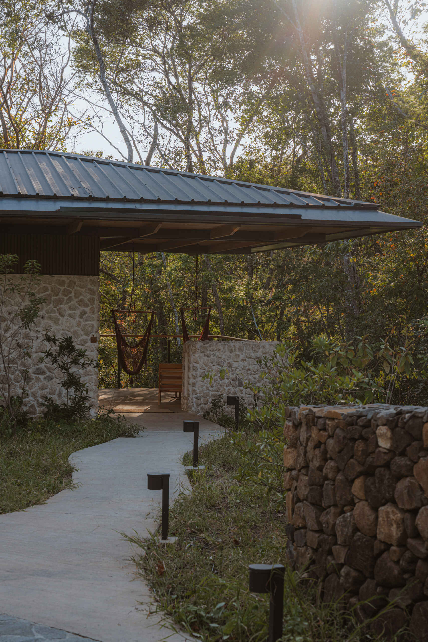 A stone-clad jungle bungalow in Costa Rica blends effortlessly into its forest surroundings. A curved concrete path leads to the entrance, while a floating terrace and rich timber ceiling invite guests into a space that feels both grounded and elevated.