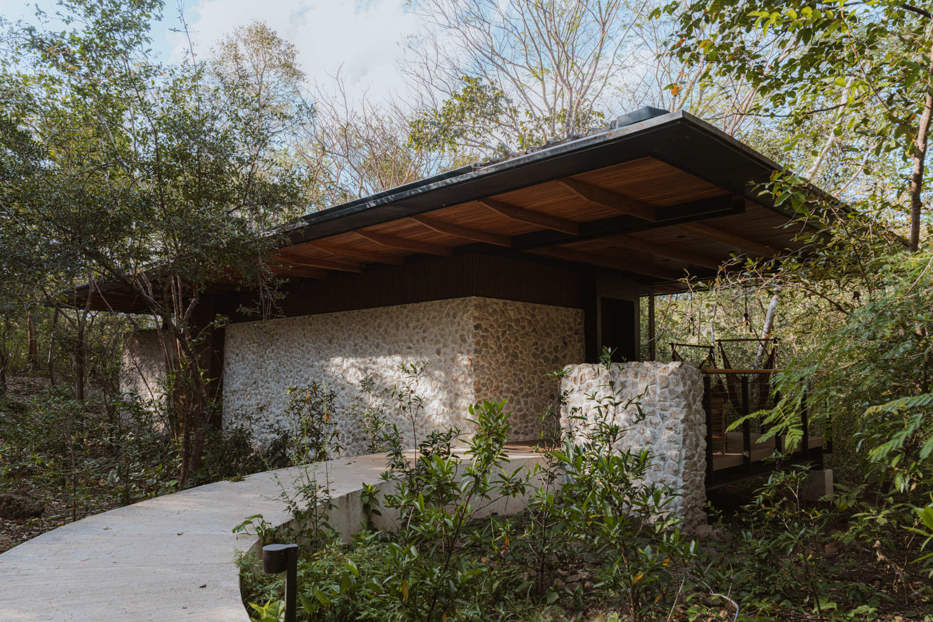 A stone-clad jungle bungalow in Costa Rica blends effortlessly into its forest surroundings. A curved concrete path leads to the entrance, while a floating terrace and rich timber ceiling invite guests into a space that feels both grounded and elevated.