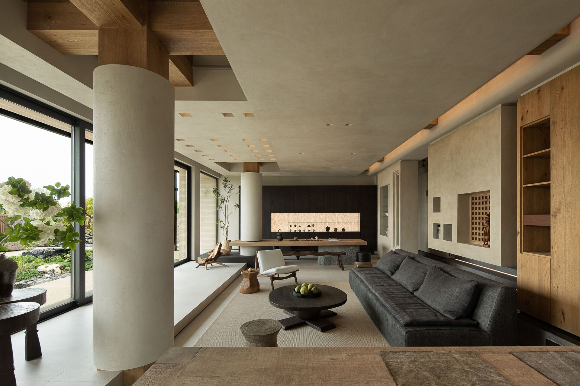 The dark wood cabinetry at the end of the living room sets a moody backdrop to the large wood table, while a softly lit niche displays delicate ceramics like a personal gallery. Every element, from the curved bench seat to the tall potted branch, feels thoughtfully placed, creating a room that’s serene, grounded, and full of quiet soul.