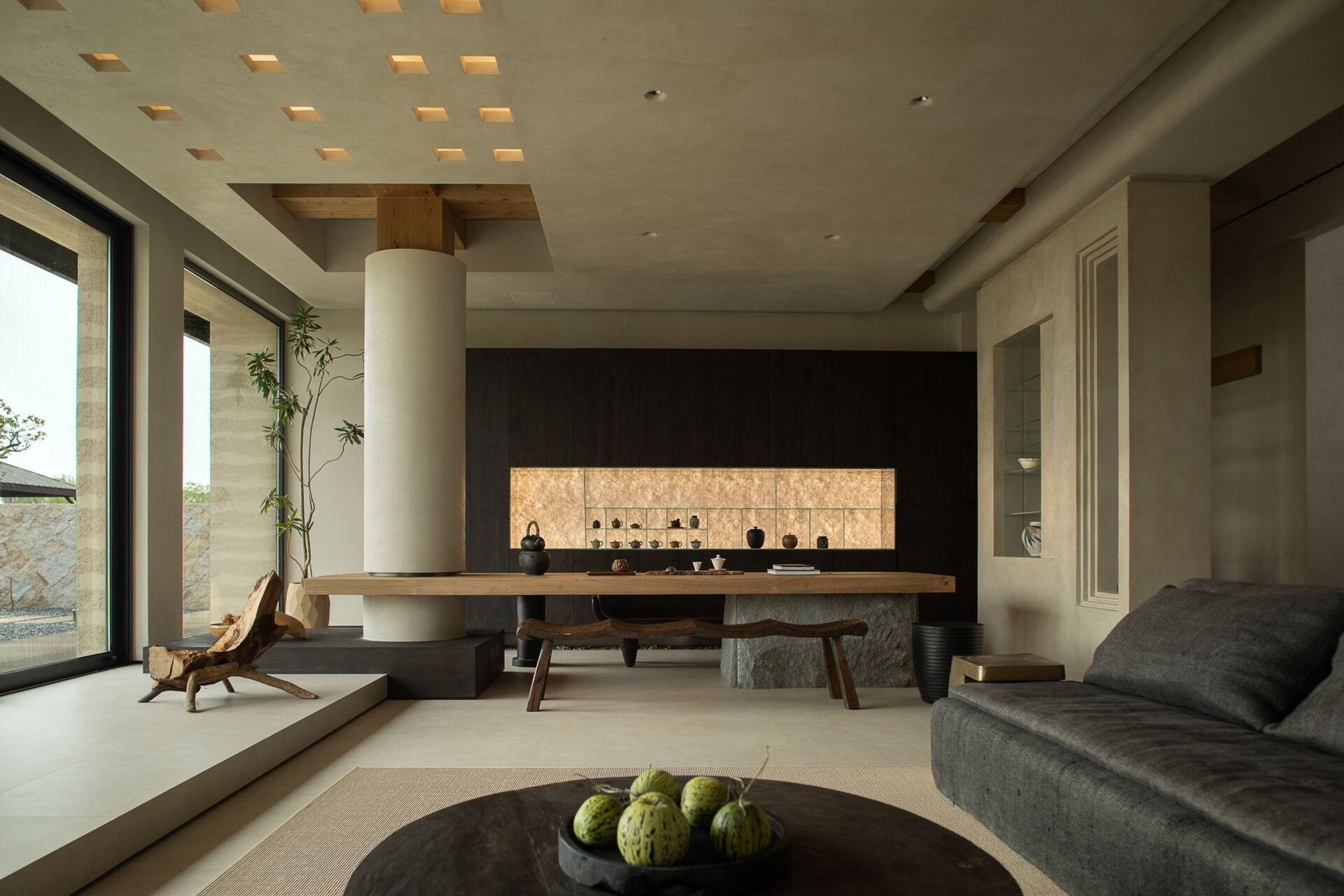 The dark wood cabinetry at the end of the living room sets a moody backdrop to the large wood table, while a softly lit niche displays delicate ceramics like a personal gallery. Every element, from the curved bench seat to the tall potted branch, feels thoughtfully placed, creating a room that’s serene, grounded, and full of quiet soul.