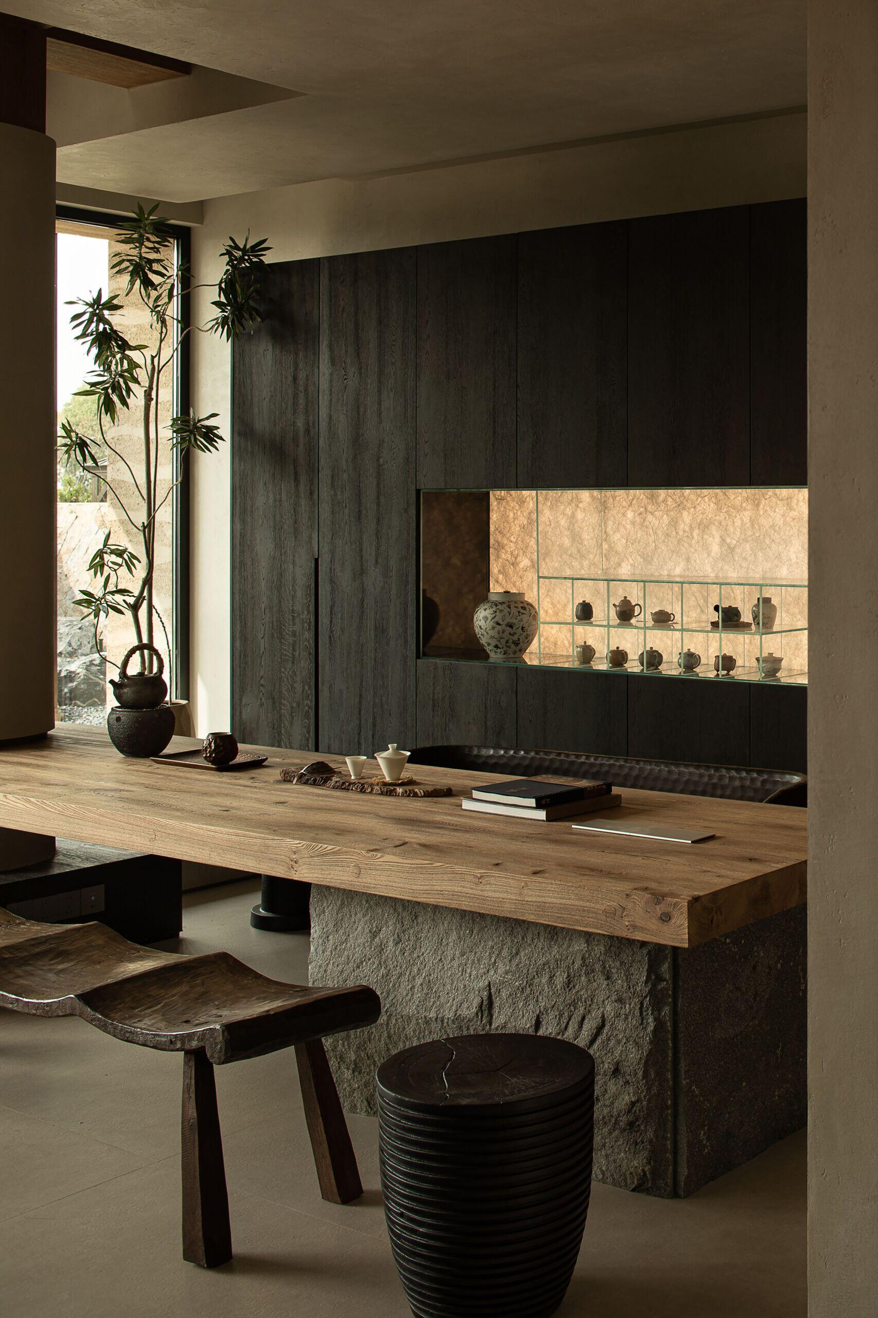 The dark wood cabinetry at the end of the living room sets a moody backdrop to the large wood table, while a softly lit niche displays delicate ceramics like a personal gallery. Every element, from the curved bench seat to the tall potted branch, feels thoughtfully placed, creating a room that’s serene, grounded, and full of quiet soul.
