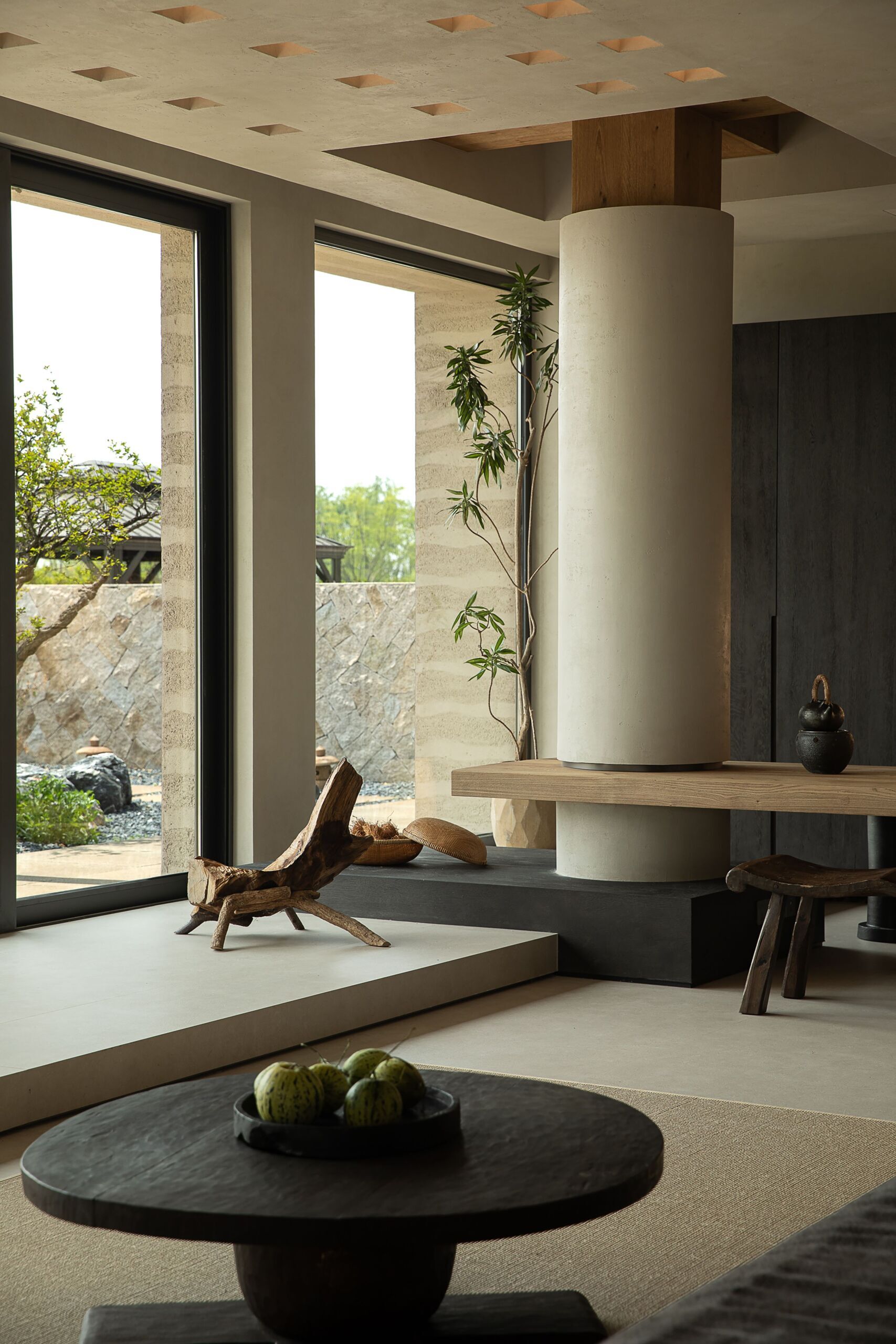 The dark wood cabinetry at the end of the living room sets a moody backdrop to the large wood table, while a softly lit niche displays delicate ceramics like a personal gallery. Every element, from the curved bench seat to the tall potted branch, feels thoughtfully placed, creating a room that’s serene, grounded, and full of quiet soul.