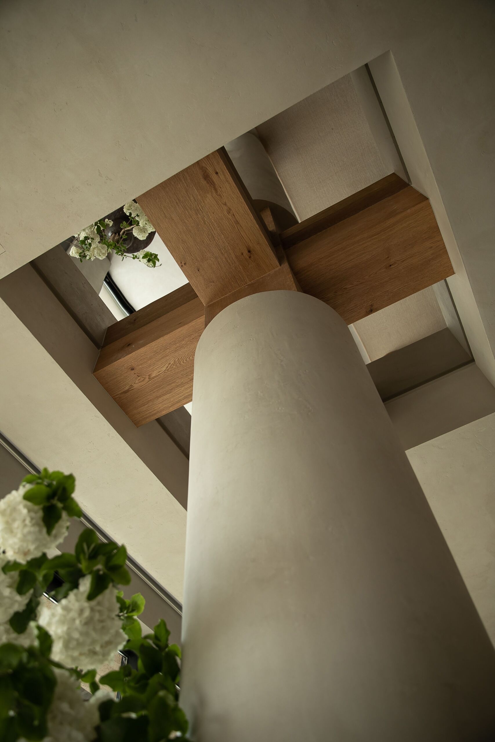 Seen from below, this sculptural column makes a striking impression with its smooth plaster surface rising to meet a bold cross of natural wood beams. The contrast between soft curves and strong lines gives the space both strength and elegance. Subtle textures in the ceiling and surrounding structure add depth, while glimpses of fresh white blossoms in the mirror softens the view.