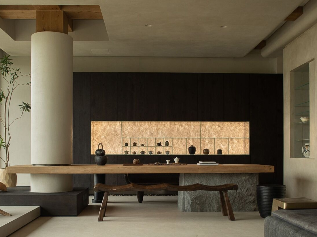 The dark wood cabinetry at the end of the living room sets a moody backdrop to the large wood table, while a softly lit niche displays delicate ceramics like a personal gallery. Every element, from the curved bench seat to the tall potted branch, feels thoughtfully placed, creating a room that’s serene, grounded, and full of quiet soul.