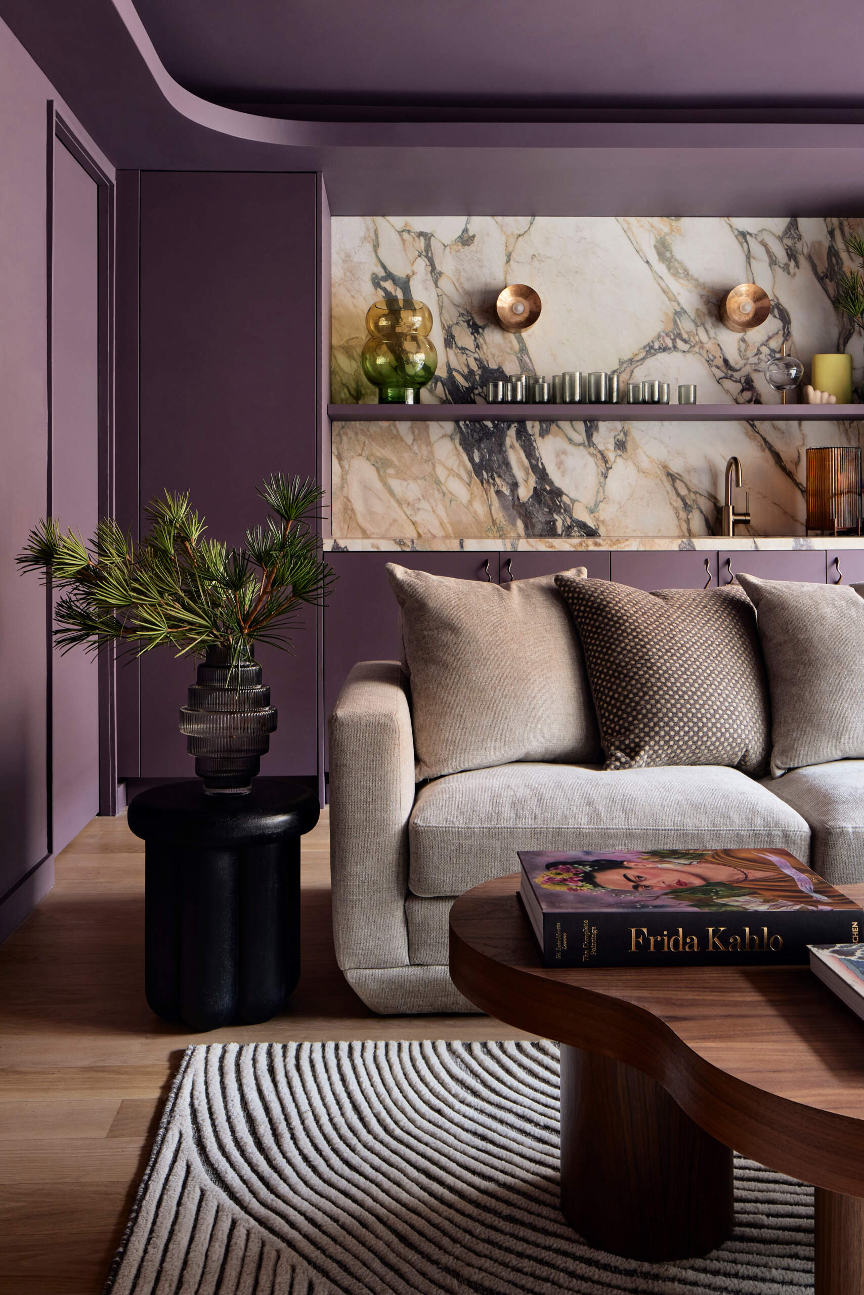 This media room, painted in deep aubergine, features a marbled bar and chartreuse velvet seating surrounding a custom walnut coffee table. Curved cove lighting and a vintage Italian resin lamp bring softness to the dramatic palette.
