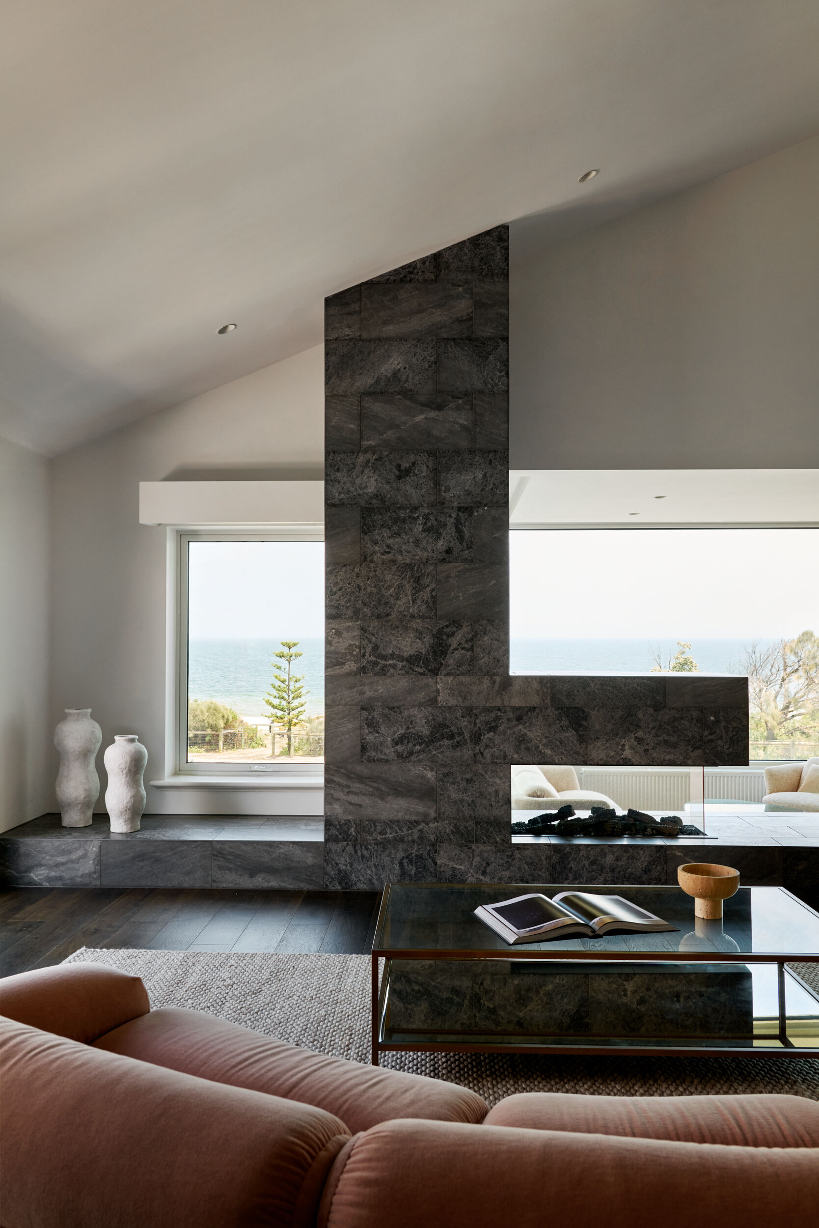 In the living room, a monolithic fireplace clad in dark stone anchors the room with its sculptural presence. The stone’s depth and texture catch the light differently throughout the day, adding quiet movement to an otherwise solid form. A peek-through opening in the fireplace creates a subtle connection to the adjacent lounge while keeping the atmosphere cozy and contained. In front, a plush rose-colored sofa softens the space with gentle curves and a velvety texture.