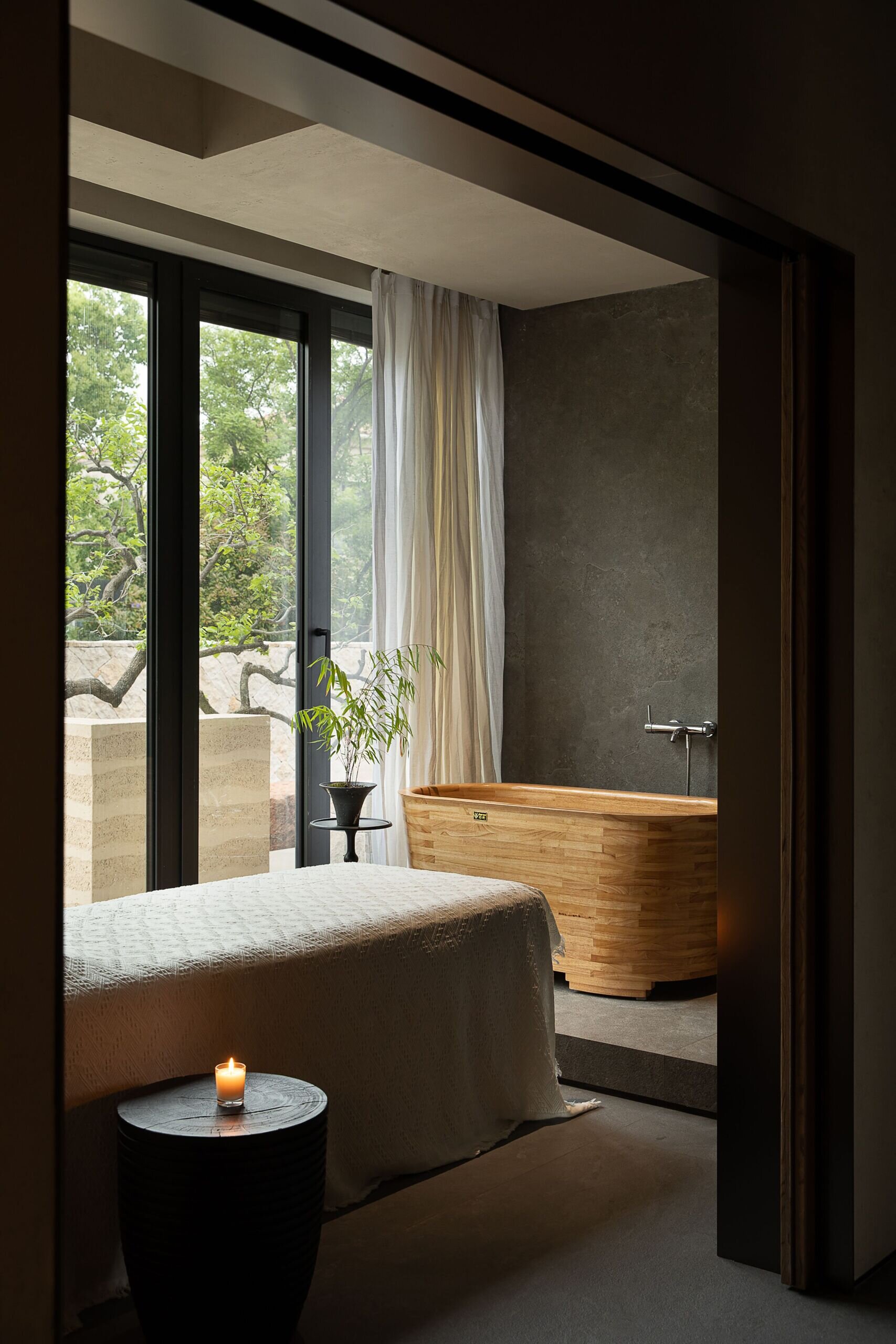 This tranquil massage room feels like a personal retreat designed for total relaxation. A soft white cover drapes the massage table, while a small flickering candle adds a gentle glow to the earthy palette. The wooden soaking tub brings warmth and craft to the space, its curved lines inviting you to slow down and breathe.