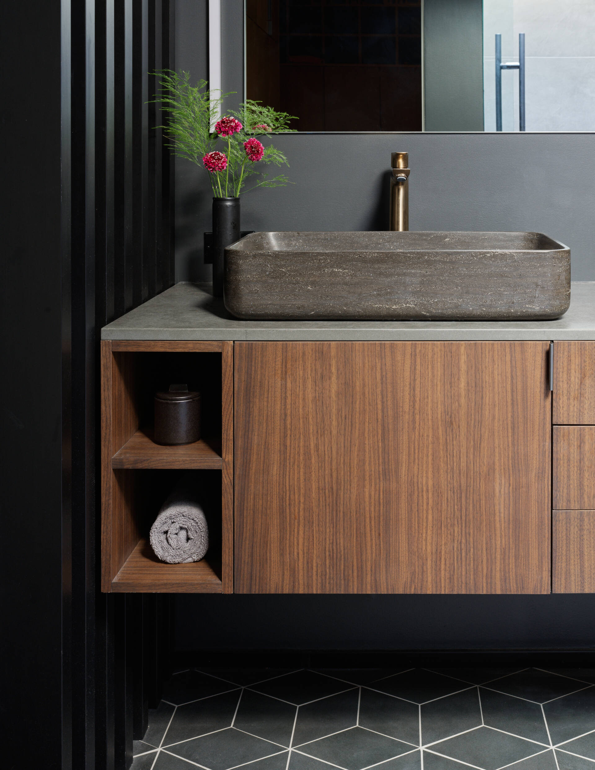 This Mid-Century Modern inspired bathroom vanity pairs rich walnut cabinetry with bold geometric tile and a sculptural stone sink. Brushed bronze accents and a sleek black screen wall add warmth and drama to the serene, minimalist design.