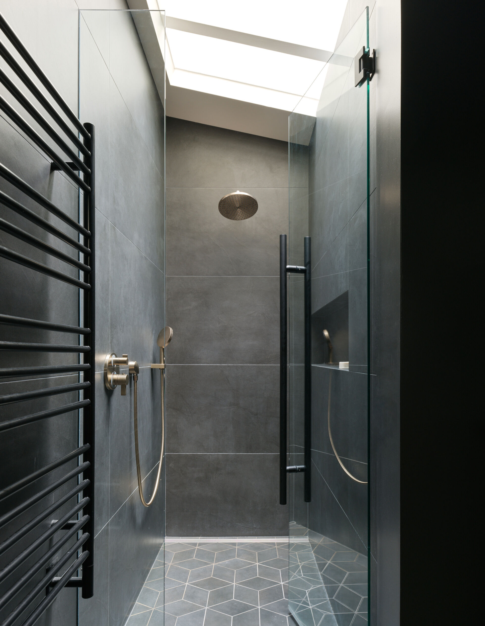 This shower pairs dark tile with brushed bronze fixtures and a frameless glass door for a clean, architectural look. A skylight above fills the space with natural light, turning the shower into a calming, spa-like retreat.