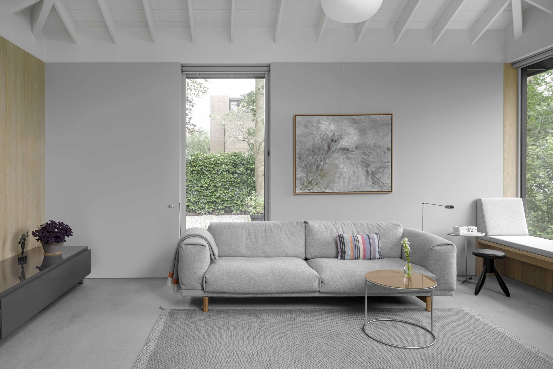 This living room continues the visual rhythm of oak, concrete, and light. A built-in window seat beneath expansive glazing offers a peaceful corner to read, rest, or enjoy time with grandchildren. The exposed rafters above enhance the architectural openness, giving this quiet space a soaring, unpretentious character.