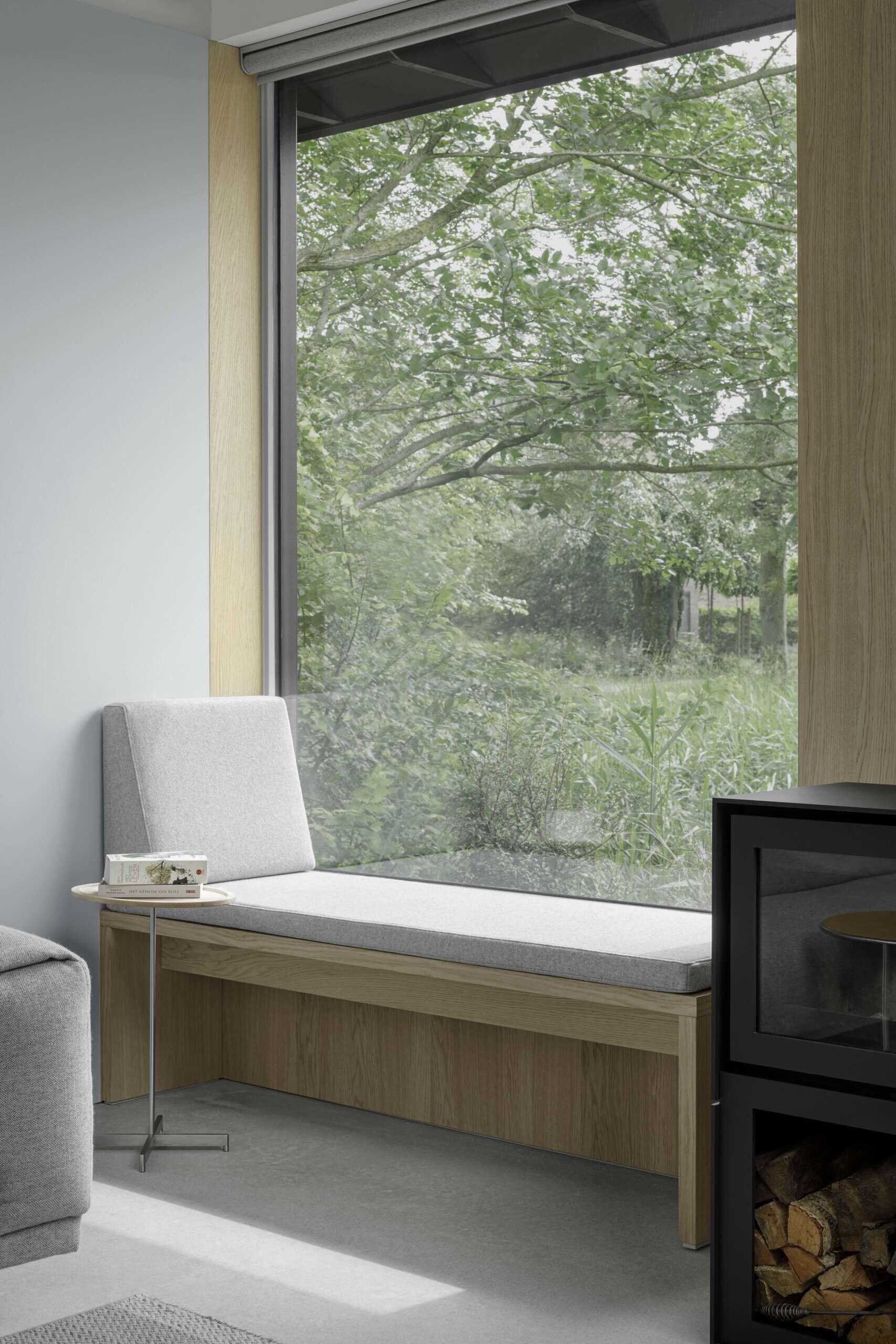 This living room continues the visual rhythm of oak, concrete, and light. A built-in window seat beneath expansive glazing offers a peaceful corner to read, rest, or enjoy time with grandchildren. The exposed rafters above enhance the architectural openness, giving this quiet space a soaring, unpretentious character.