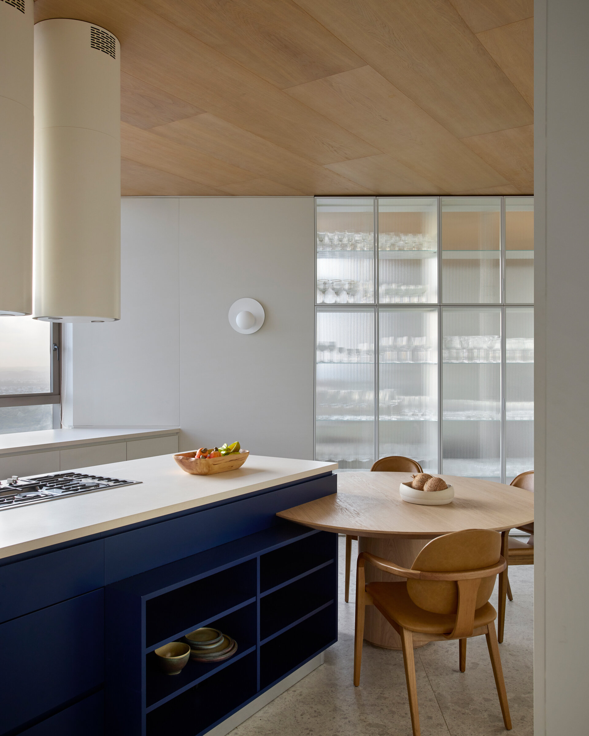 The kitchen and barbecue area share the upper floor with the main social spaces. The kitchen features a countertop clad natural stone, while the blue cabinetry echoes the tones of the sea. Materials and layout of the barbecue area emphasize openness and continuity, with finishes chosen to unify the communal areas.