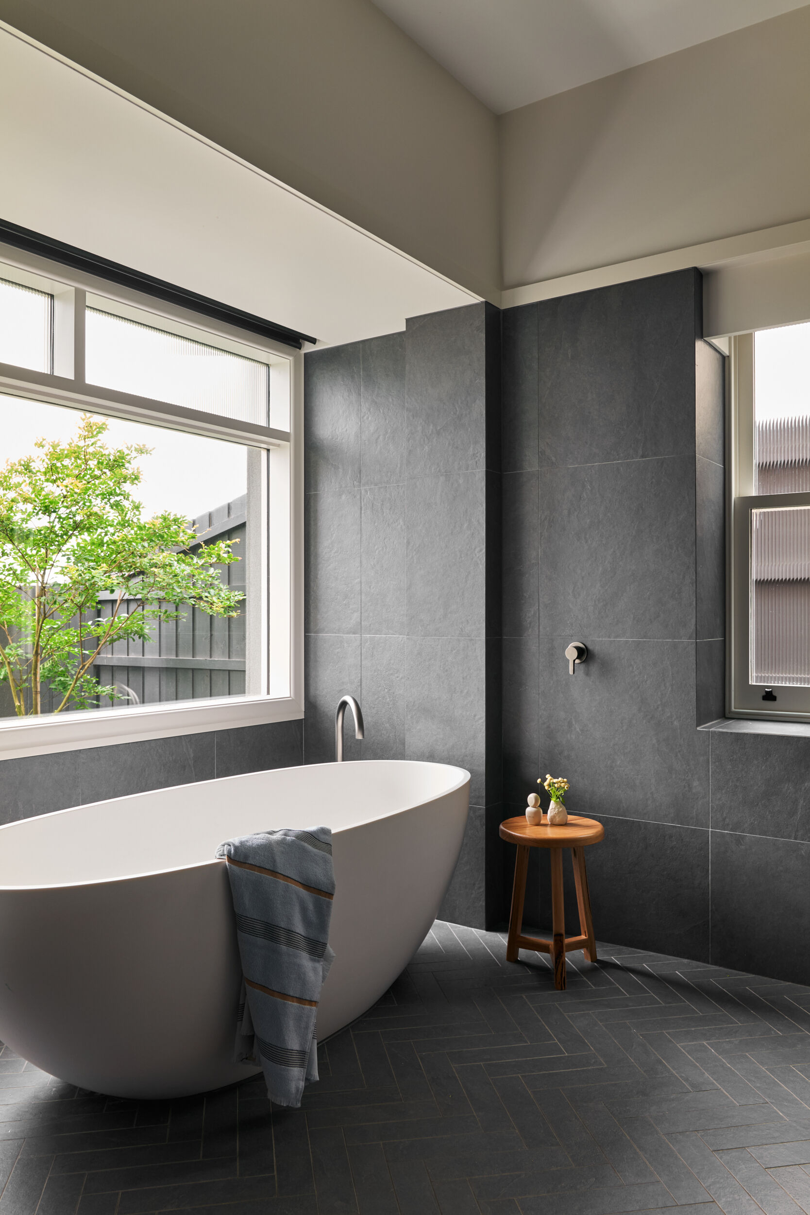 Bathed in soft natural light, this bathroom feels like a personal spa retreat—quiet, minimal, and deeply calming. The centerpiece is a sculptural freestanding tub, its smooth matte surface a gentle contrast against the textured dark tile walls that wrap around the space like a protective shell. The herringbone floor introduces subtle pattern underfoot, adding just enough rhythm to keep the design feeling grounded and intentional. A slender metal spout arcs gracefully over the edge of the tub, its simplicity adding to the overall sense of ease.