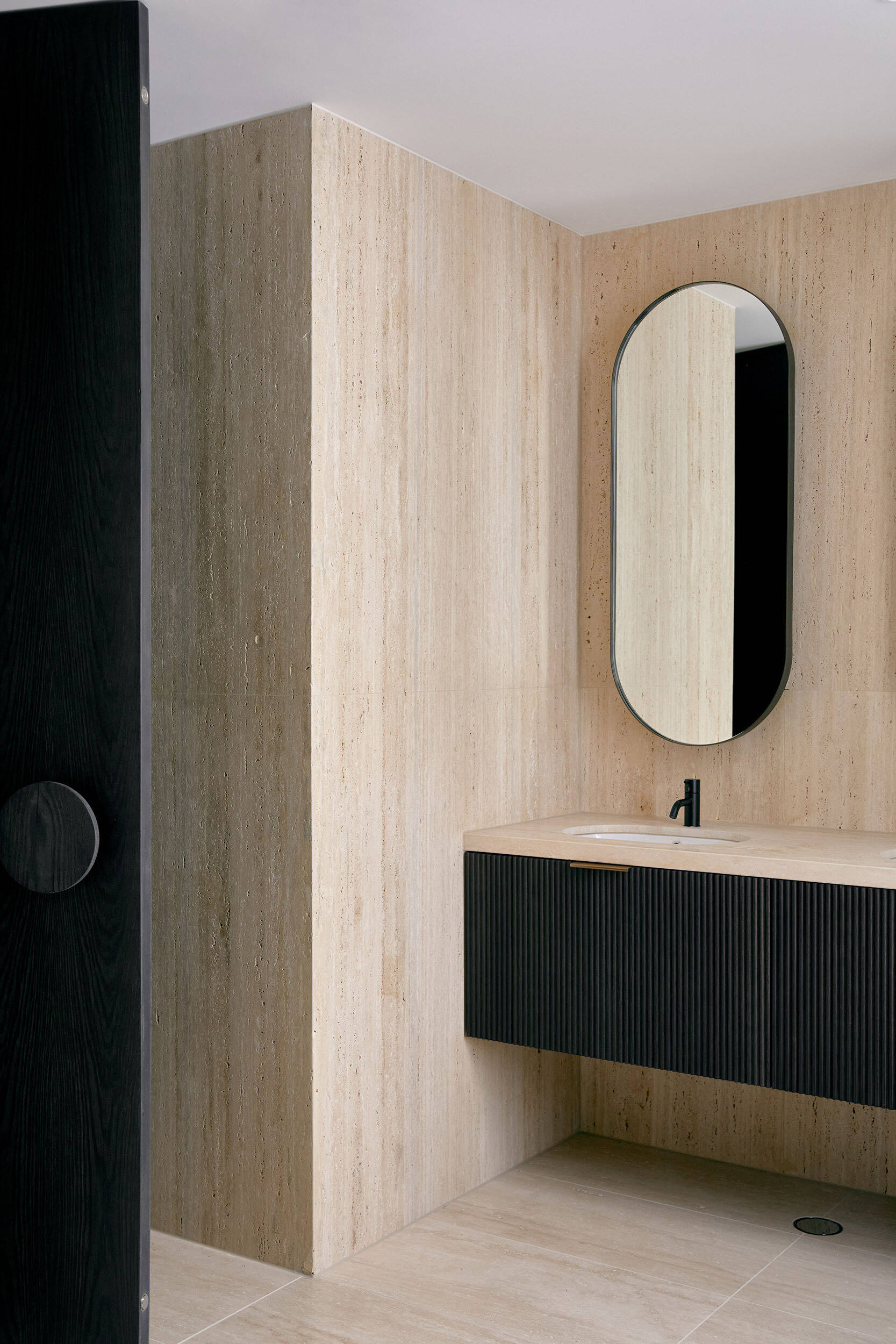 In this bathroom, warm travertine tiles wrap the floor and walls, creating a calm, monolithic backdrop. A fluted black vanity floats effortlessly above the ground, paired with a pale stone benchtop that softens the contrast. Details are restrained but deliberate, from the matte black tapware to the pill-shaped mirror that echoes the project’s sculptural curves.