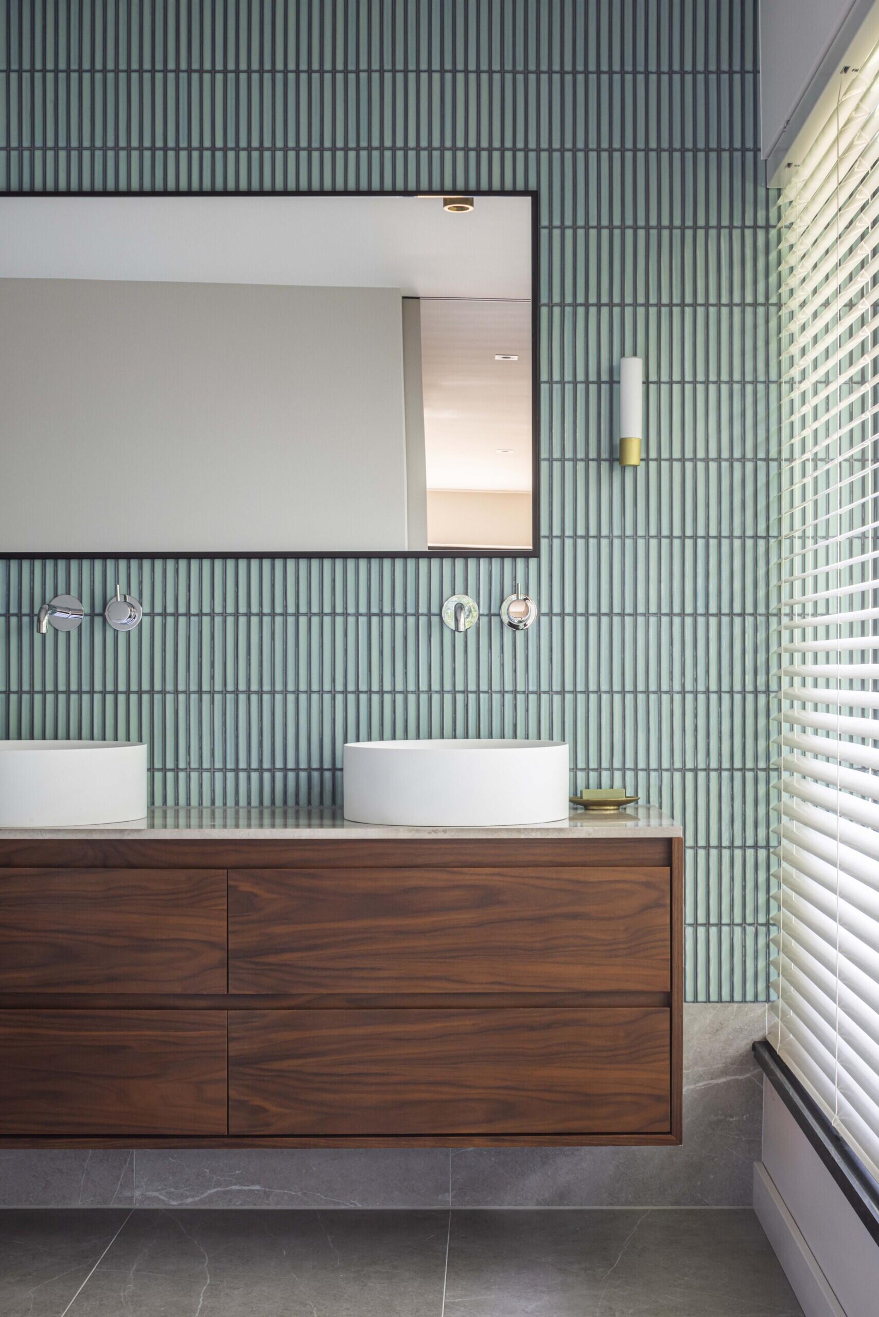 This serene bathroom pairs refined materiality with clean geometry. Vertical blue tiles form a subtle rhythm across the wall, grounding the space in texture and calm. A floating walnut vanity adds warmth beneath dual vessel sinks, while the oversized mirror and minimalist fixtures contribute to the room’s quiet sophistication. Gentle natural light filters through blinds, softening the bold contrast of stone, wood, and ceramic.