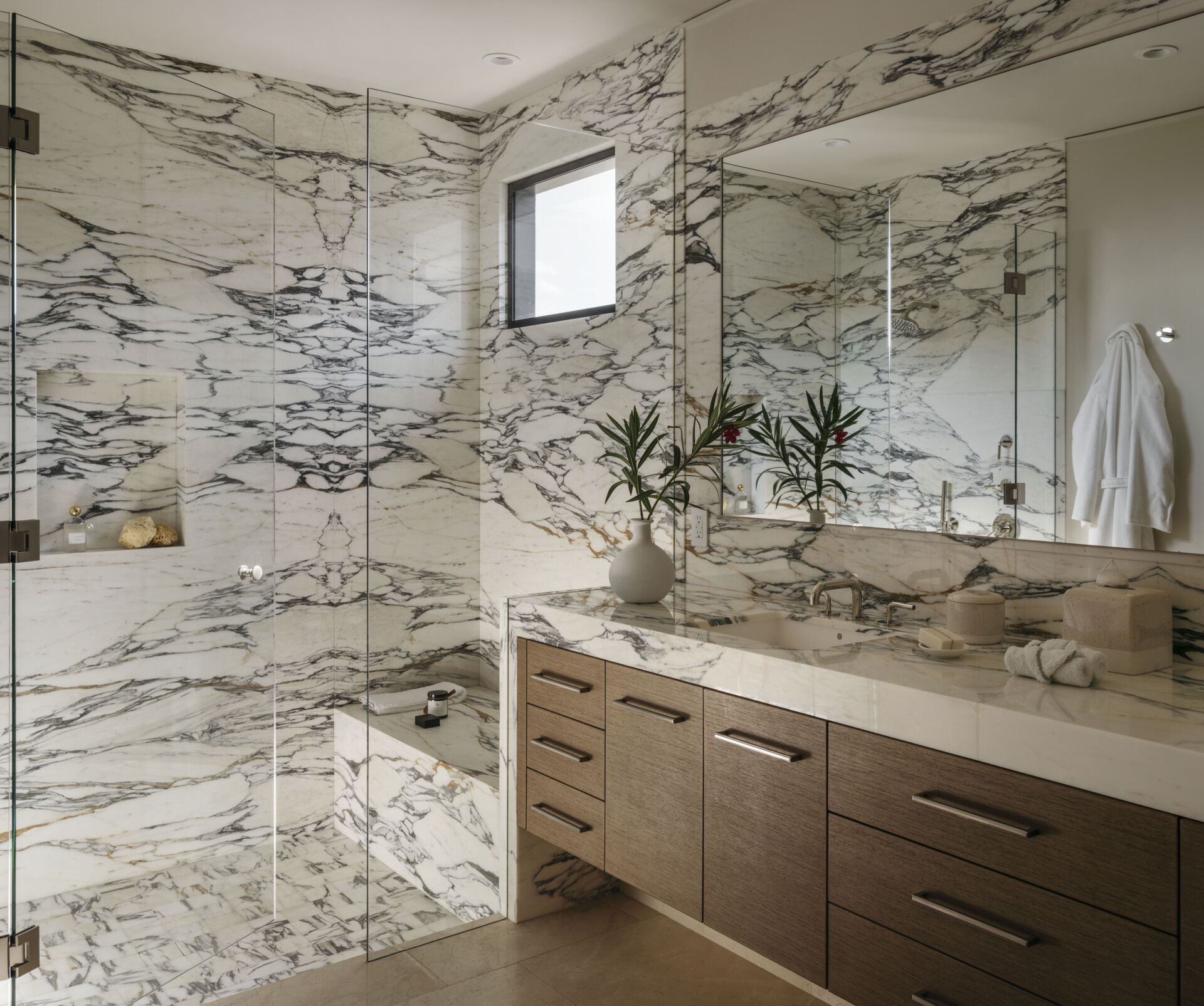 This bathroom feels like a private retreat carved from a single slab of stone. The dramatic veining of the marble wraps continuously across walls, shower, and vanity, creating a visual rhythm that’s both bold and serene.
The floating vanity, with its understated wood grain drawers and sleek metal pulls, adds a gentle contrast, grounding the space without disrupting its flow.
Even the built-in shower bench and recessed shelf seem thoughtfully integrated, offering function without fuss.