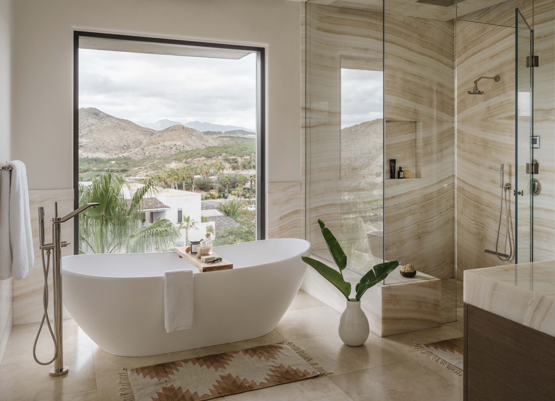This bathroom has a sculptural tub sits confidently in front of a wide picture window, framing rolling desert hills and lush palms below. Every surface inside plays with warm, sandy tones, from the smooth stone floor to the gently striated walls that echo the landscape outside. The walk-in shower is wrapped in the same soft stone, creating a sense of continuity that feels calming and intentional.