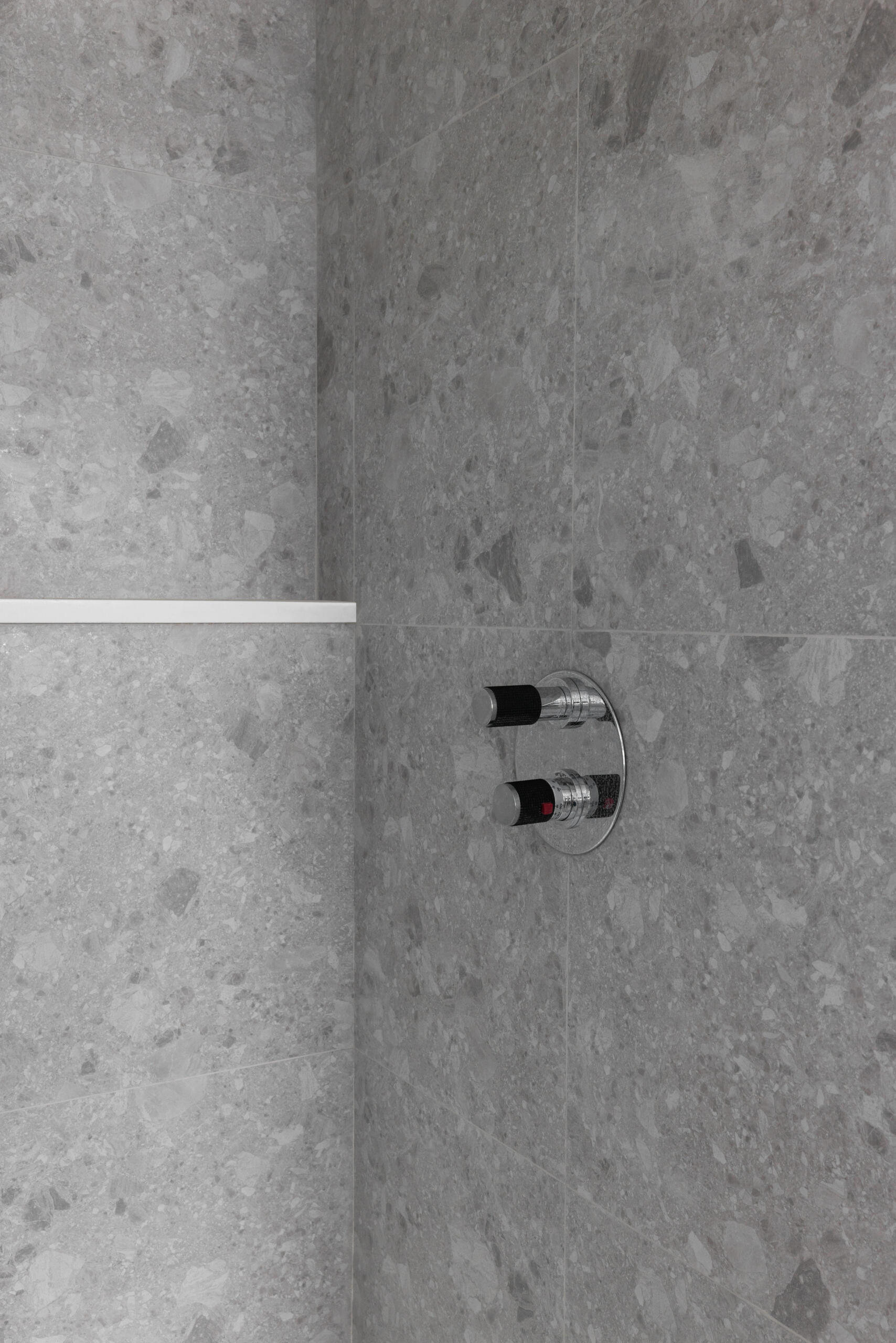 This close-up of the shower captures a quiet kind of luxury, one that’s rooted in simplicity and texture. The stone tiles, full of natural variation and soft gray tones, create a sense of calm enclosure, like a modern cave made for unwinding. A thin white ledge breaks the pattern just enough to draw the eye, offering a clean place for daily essentials. The chrome controls, set flush against the wall, keep the look minimal but practical. It’s the kind of detail that makes the whole space feel intentional and thoughtfully designed, even in its quietest corners.