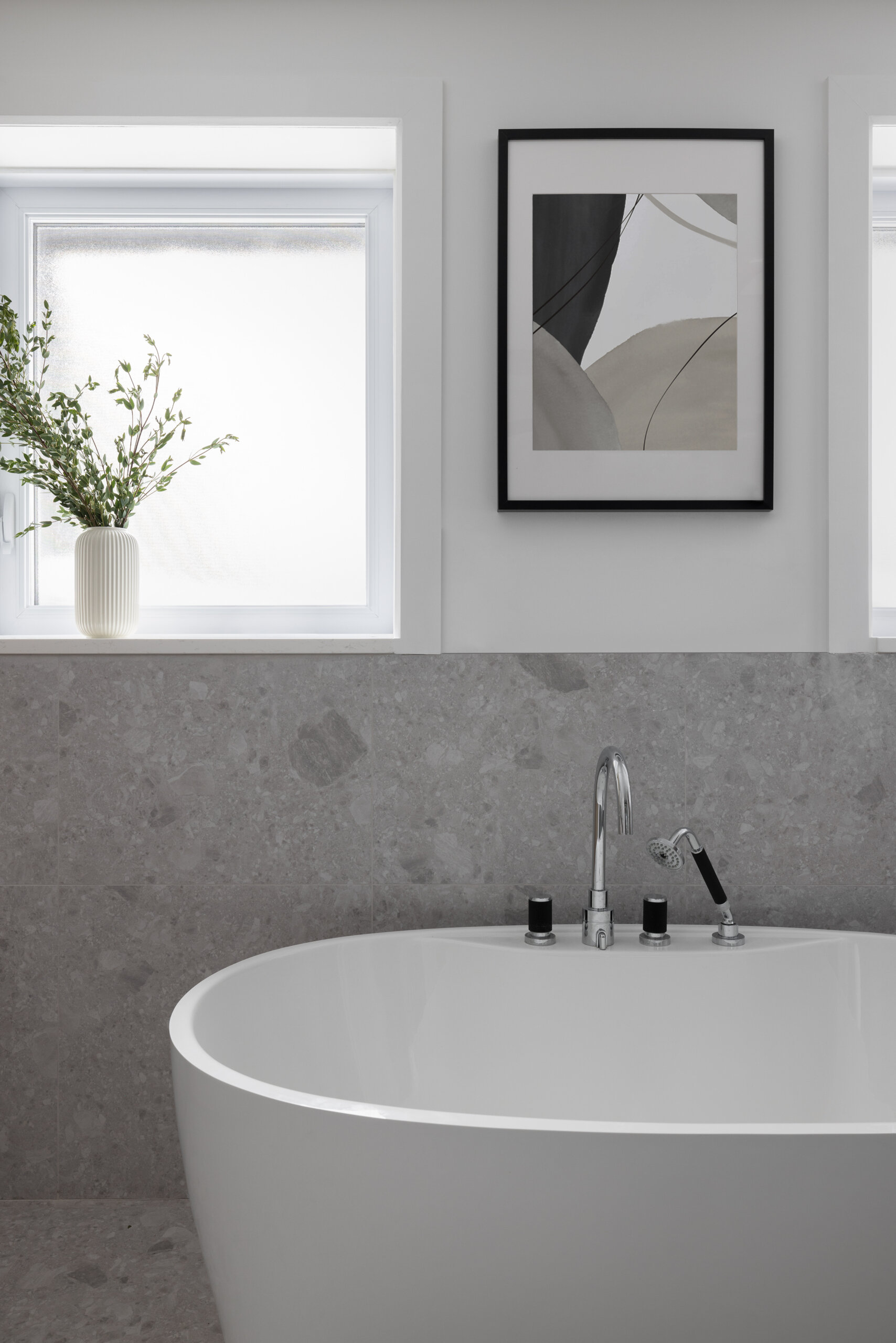 There’s something deeply soothing about this bathroom scene. The freestanding tub curves gently against a wall of soft gray stone, inviting long, unhurried soaks. Frosted windows let in plenty of daylight while keeping things private, giving the space a bright but quiet feel. A simple black-and-white abstract print adds just enough contrast, and the ribbed vase with fresh greenery brings a soft, organic touch. With its muted palette and uncluttered surfaces, this is the kind of spot that encourages you to breathe a little slower and linger just a little longer.