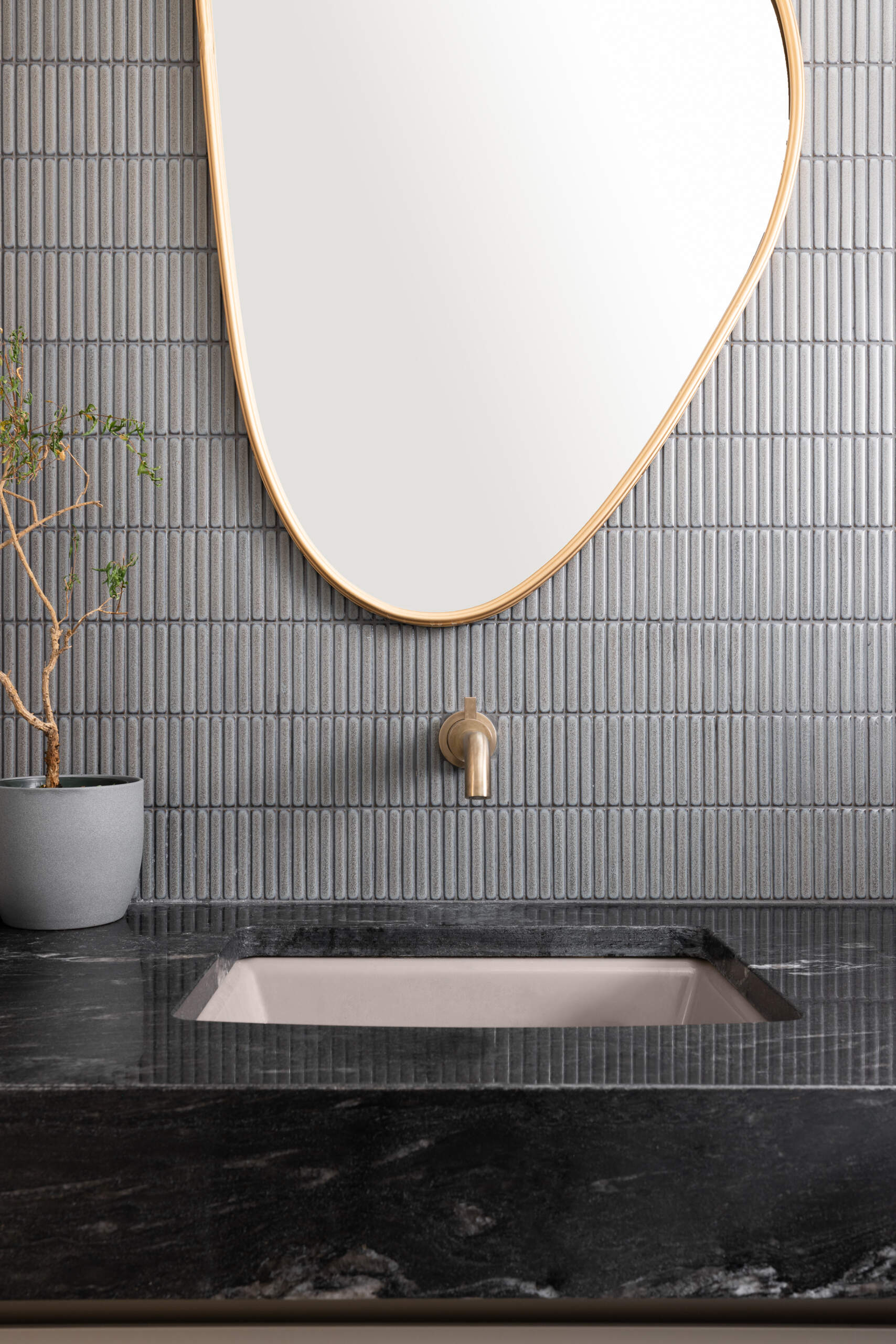 This bathroom vanity strikes a beautiful balance between organic calm and sculptural precision. The soft curves of the mirror bring warmth to the cool, vertically ridged tile, while the matte brass faucet adds a quiet touch of elegance. Anchoring it all is the dark stone countertop, its subtle veining giving a sense of depth and natural movement. A single potted plant adds a breath of life to the space, inviting a slower, more mindful pace. It’s the kind of setting that makes even a quick rinse feel like a moment of calm in the middle of a busy day.