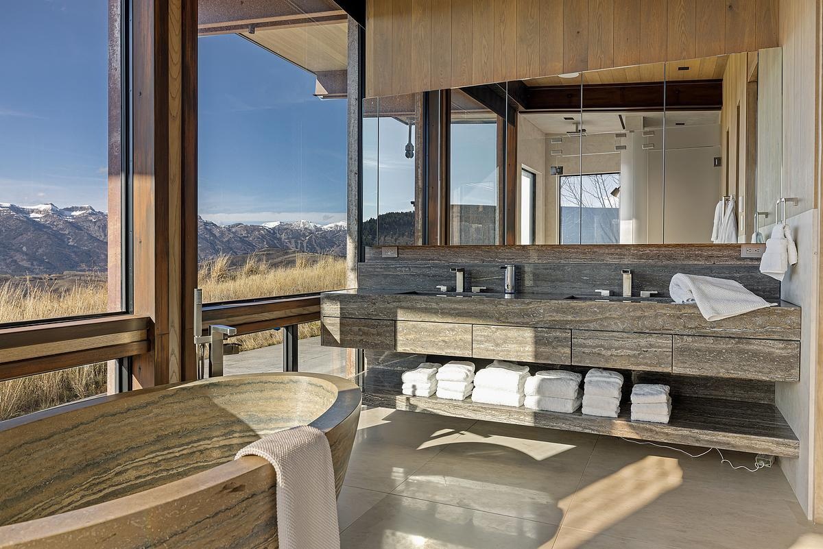 Soaked in natural light and surrounded by mountain views, this bathroom feels more like a spa tucked into the hillside. The stone tub, with its smooth curves and layered texture, invites long, restorative soaks with nature as the backdrop. A floating vanity in rich, veined stone adds a sleek, grounded feel, while open shelving keeps plush towels within reach. With warm wood details and expansive glass, the space balances raw beauty with refined calm, a place to reset, breathe deep, and simply be.