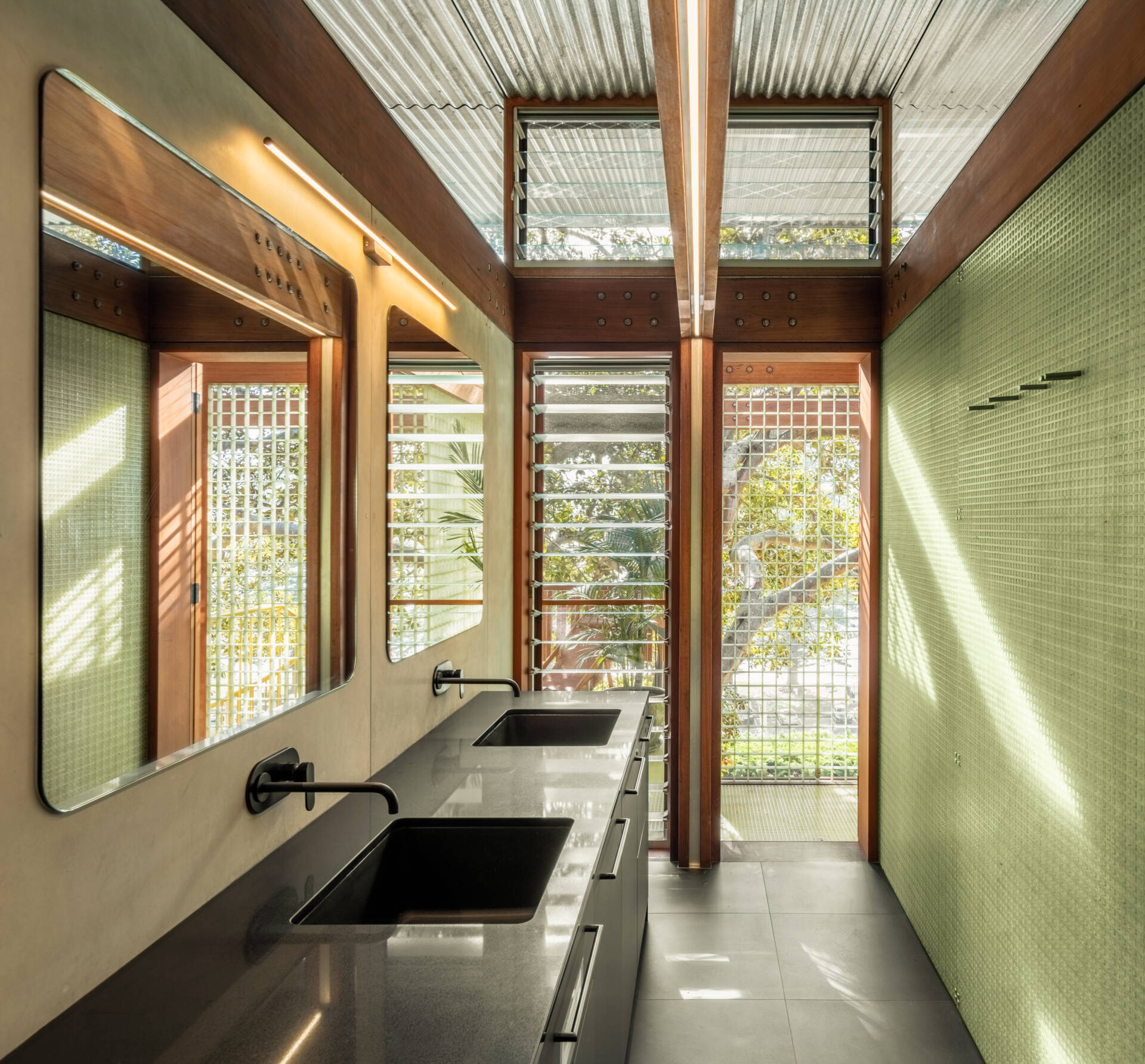 Clad in soft green tiles and wrapped in timber, the bathroom feels fresh and unfussy. Matte black tapware and a dark vanity add contrast without fuss, while upper glazing and slatted windows bring in dappled light from the canopy.