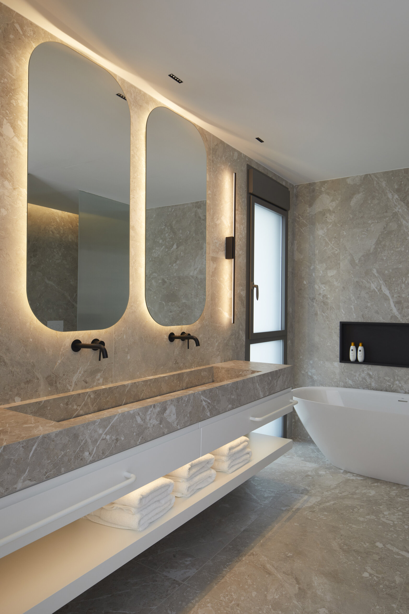 In the primary ensuite, a long stone vanity anchors this calming space, paired with softly glowing mirrors and matte black fixtures for contrast. Open shelves hold neatly folded towels, while a freestanding tub and black-lined recessed niche invite quiet, unhurried routines. Wrapped in soft gray stone, the shower pairs subtle texture with sharp black fixtures and a backlit niche. A frameless oval mirror adds a gentle curve, balancing the minimal palette with a quiet sense of care.