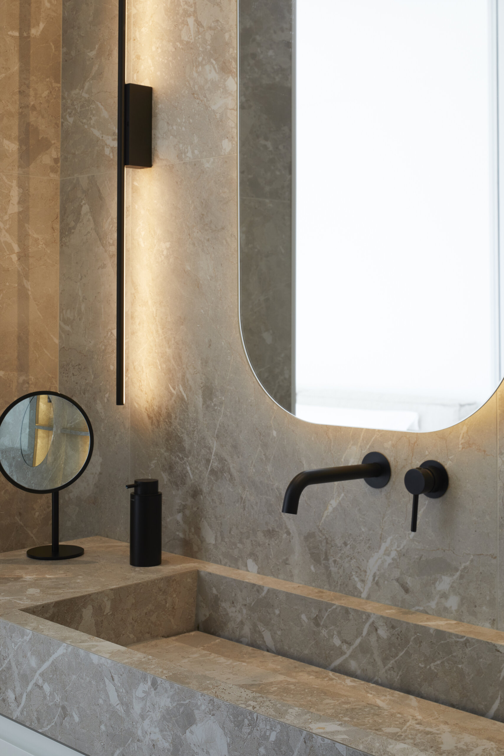 In the primary ensuite, a long stone vanity anchors this calming space, paired with softly glowing mirrors and matte black fixtures for contrast. Open shelves hold neatly folded towels, while a freestanding tub and black-lined recessed niche invite quiet, unhurried routines. Wrapped in soft gray stone, the shower pairs subtle texture with sharp black fixtures and a backlit niche. A frameless oval mirror adds a gentle curve, balancing the minimal palette with a quiet sense of care.