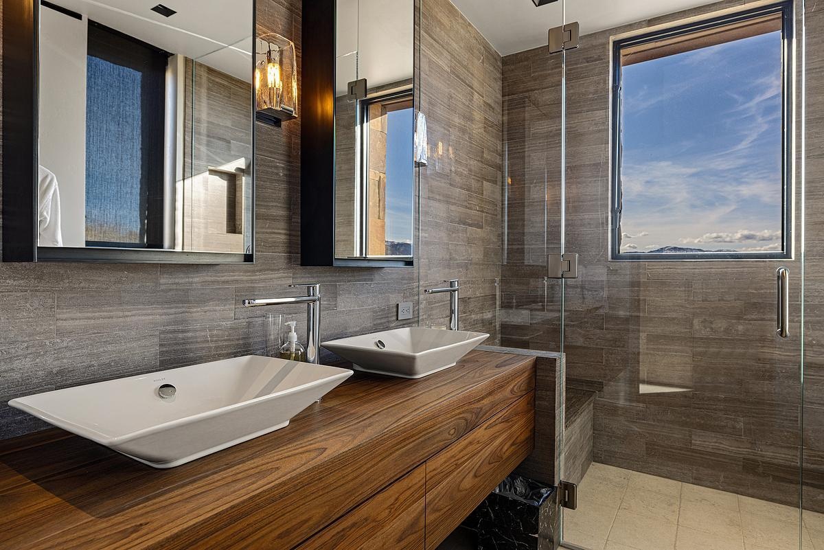 This bathroom pairs bold simplicity with warm, organic style. The wood-grain vanity brings a rich, grounded presence to the room, topped with sleek vessel sinks that feel sculptural yet unfussy. Wall-mounted mirrors and soft-glow sconces add just the right touch of refinement, while floor-to-ceiling tile in earthy gray tones ties it all together. The walk-in shower, with its frameless glass and picture-perfect window, offers a view that turns a daily ritual into something quietly luxurious.