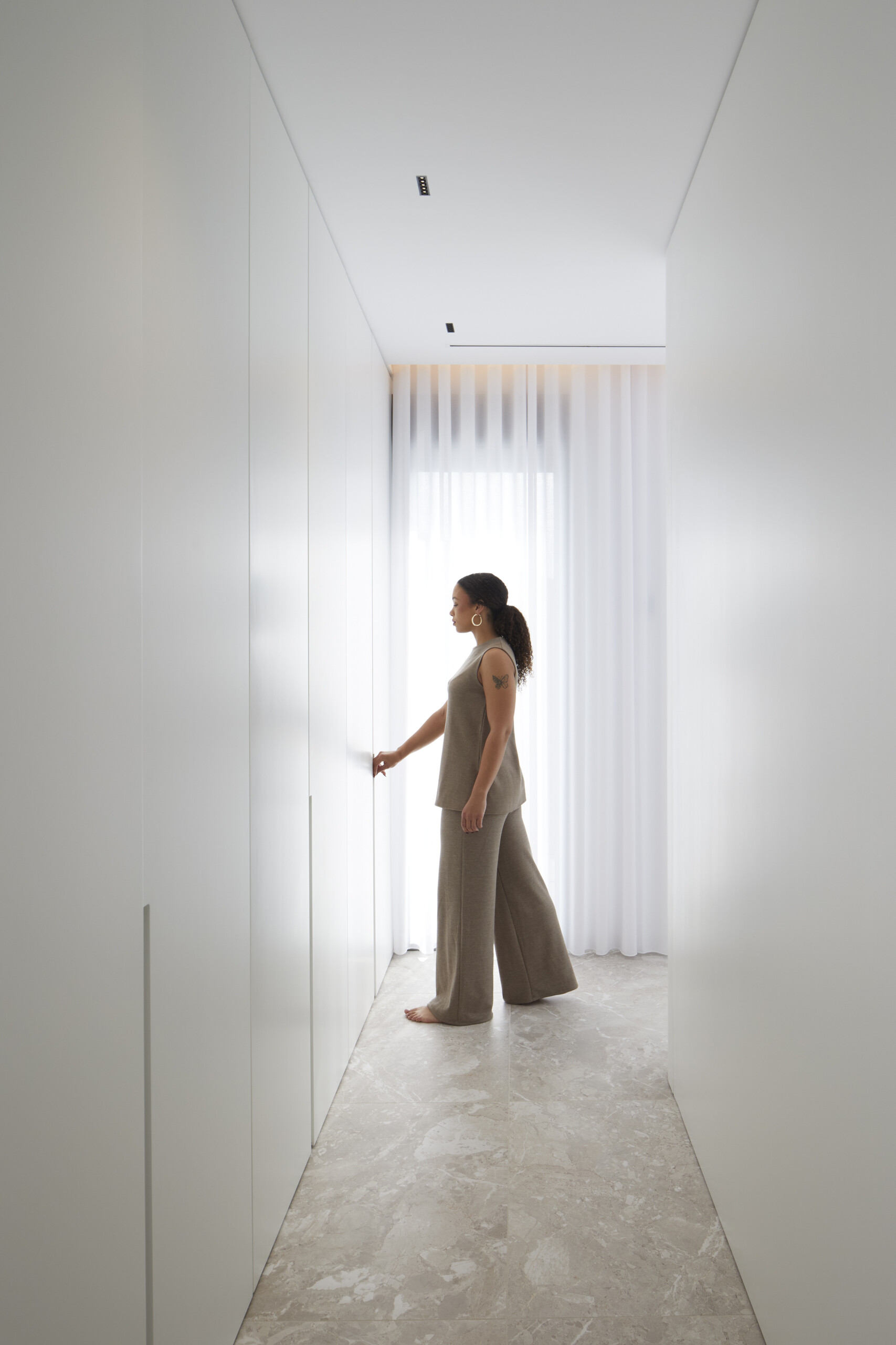 A hallway of closets reads like a gallery, flush white panels conceal generous storage, while warm stone floors and soft light at the end create a gentle, uninterrupted flow.