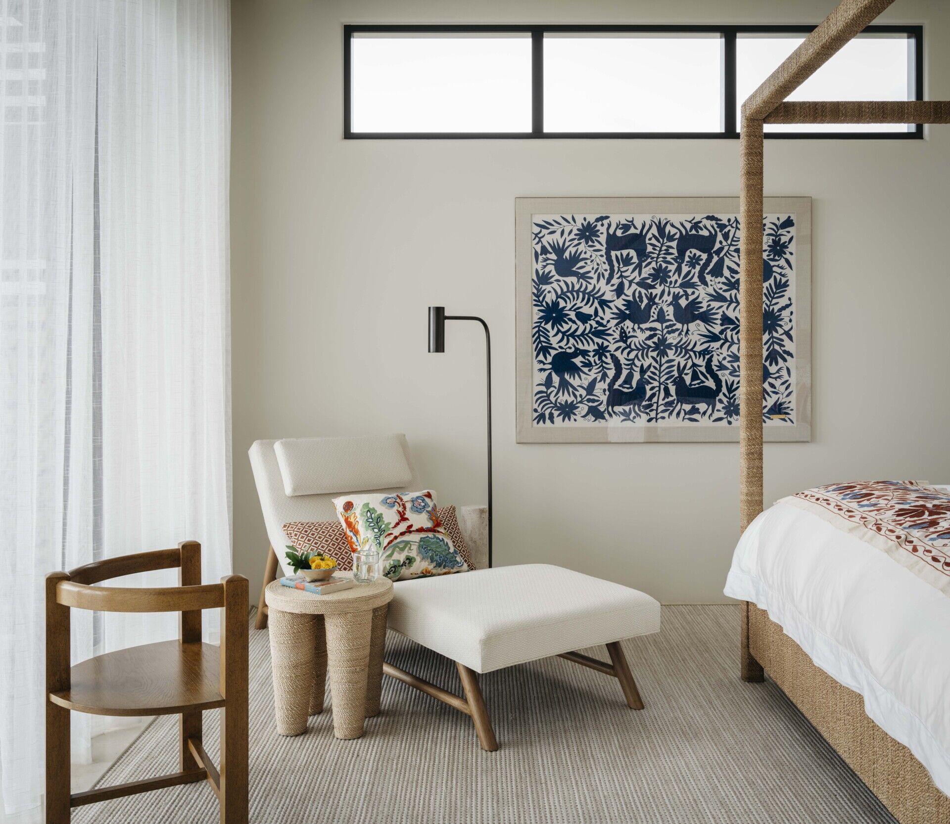 This corner of the bedroom feels like a gentle exhale. Soft natural light filters through sheer curtains, brushing over a palette of warm neutrals and inviting textures. The woven rug, the rope-wrapped bedposts, and the tactile side table all add a quiet depth without overwhelming the calm. The lounge chair, angled just so, is the perfect spot to stretch out with a book or sip something cold. Embroidered pillows bring a dash of color and personality, tying in beautifully with the deep blue artwork on the wall. That textile piece adds just enough contrast to make the room feel considered and collected.