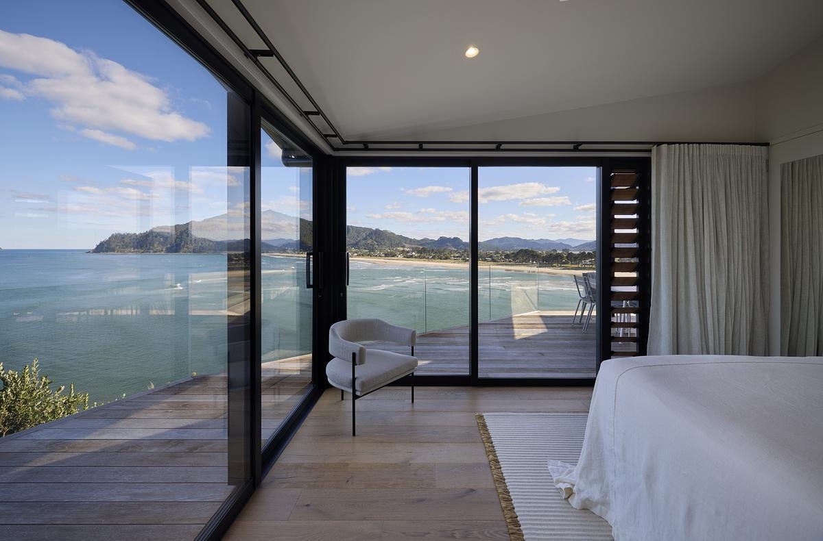 This bedroom sits just above the coastline, with floor-to-ceiling glass framing uninterrupted ocean views. Pale timber flooring and neutral-toned furnishings keep the space simple and calm, allowing the landscape to stand out. A low armchair by the window offers a spot to read or enjoy a coffee. With sliding doors that open to the breeze or close for quiet rest, the room is designed for comfort and connection to the view.