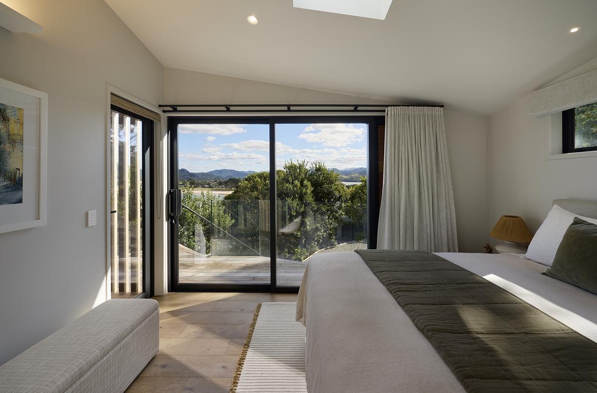 Soft light filters through sliding glass doors, opening the bedroom to views of greenery and distant hills. The space is finished in muted tones of sage, cream, and natural wood, creating a calm, cohesive feel. A textured throw and linen headboard add subtle detail, while the bench at the foot of the bed offers a practical spot to sit or take in the view. It’s a simple, restful space designed for everyday ease.