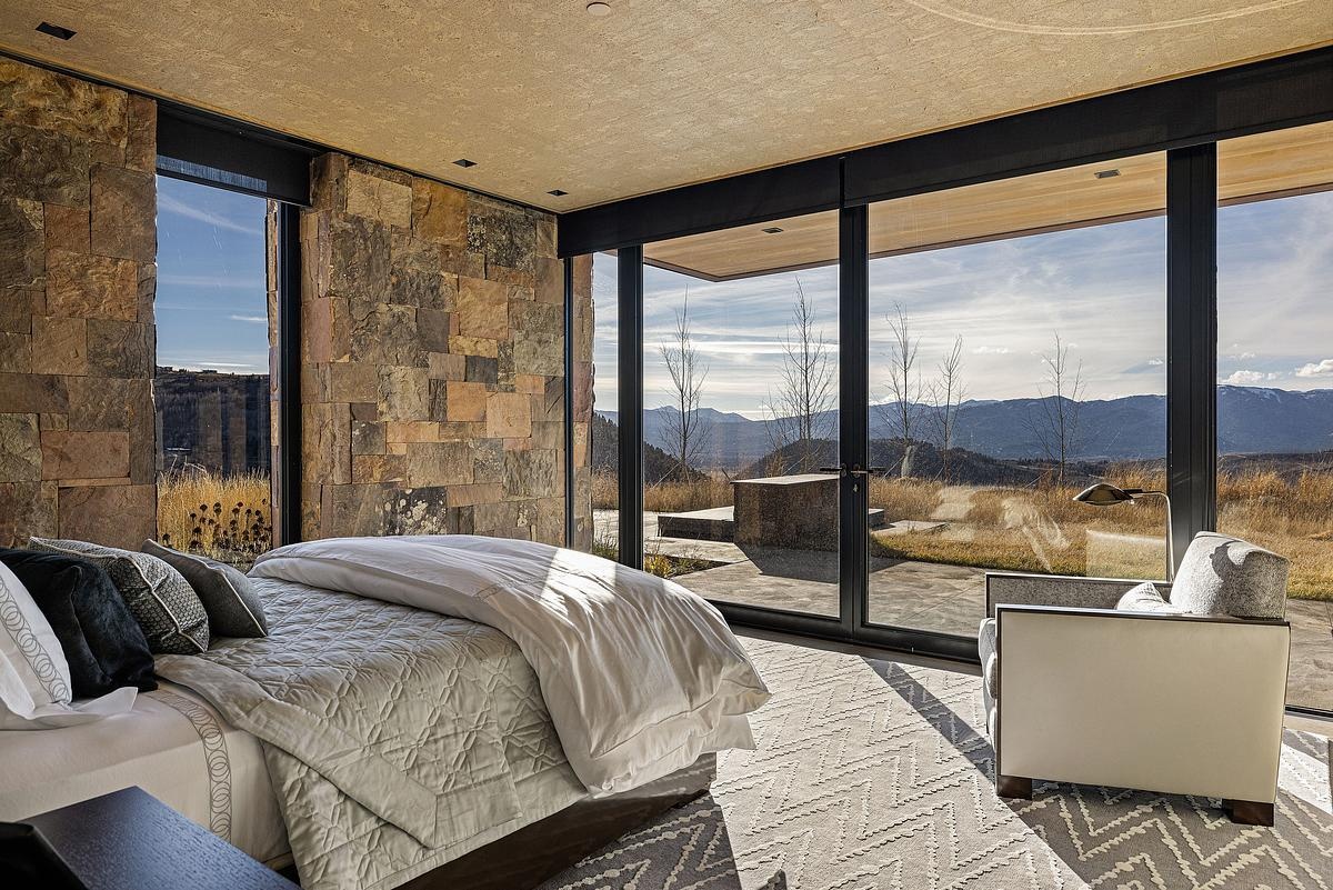 This bedroom brings the outdoors right to your bedside, with floor-to-ceiling glass offering a front-row seat to the rolling hills and morning sun. The stone walls add a natural, earthy texture that complements the muted palette of grays and creams throughout the space. Soft layers of bedding and a plush rug underfoot invite you to linger, while a cozy armchair by the window makes the perfect perch for soaking in the view. It’s a restful space that feels both grounded and expansive.